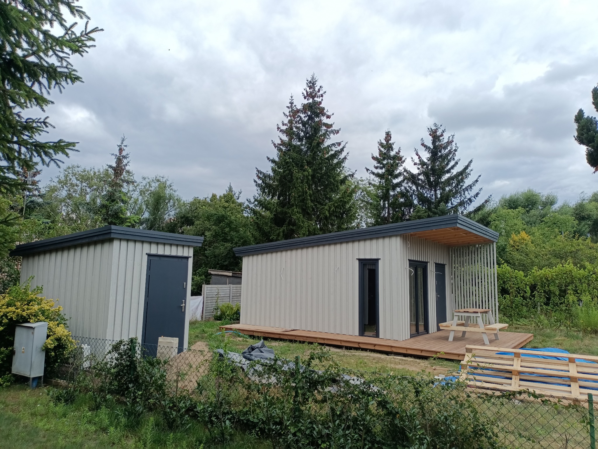 Dwa nowoczesne domki szkieletowe na działce, jeden z tarasem i stołem piknikowym. Widoczne drzewa i krzewy. Pochmurne niebo w tle. Metalowe wykończenia.