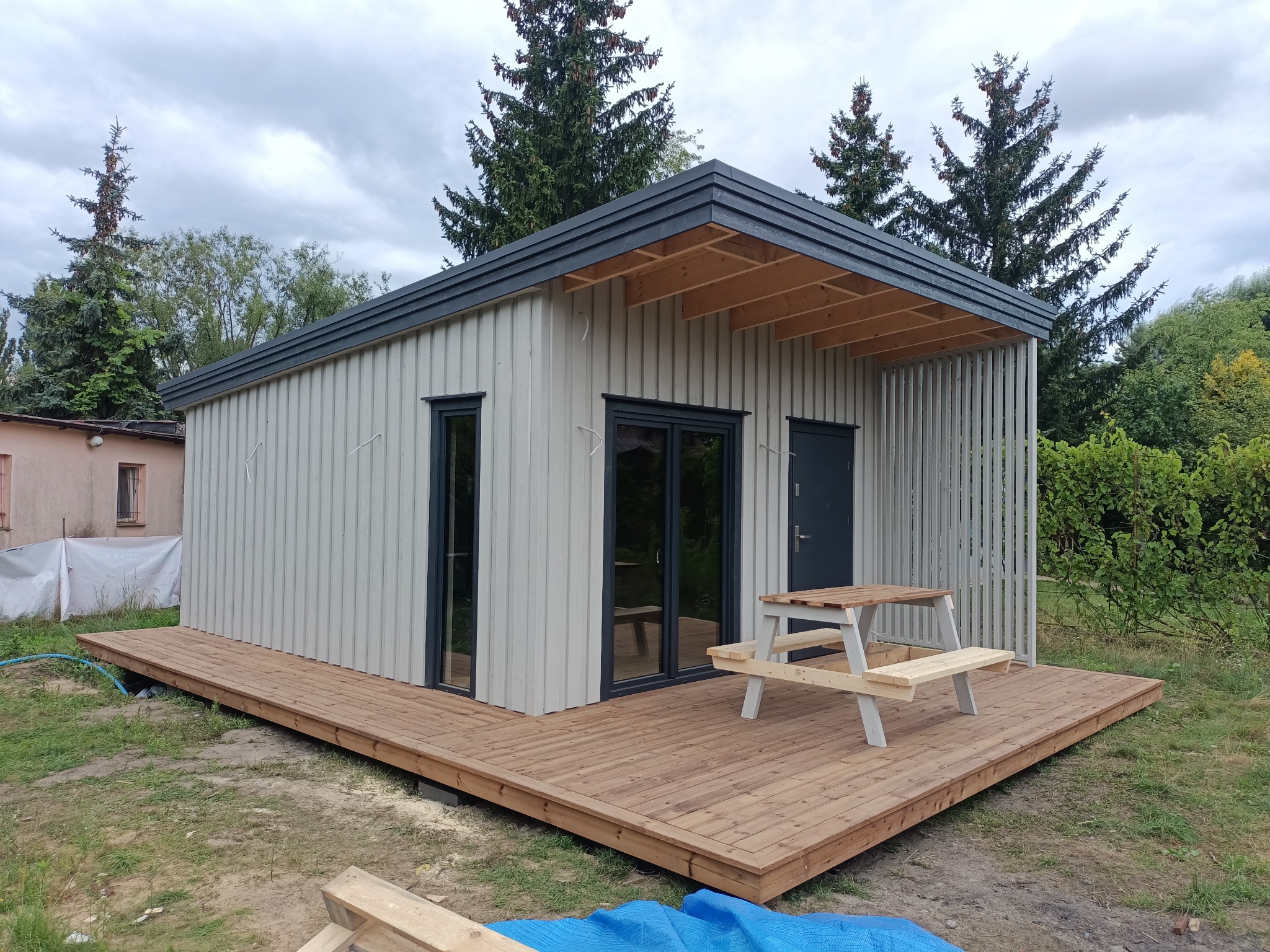 Nowoczesny, parterowy dom szkieletowy z drewnianym tarasem i stołem piknikowym. Uwagę zwraca minimalistyczna bryła i pionowe deski elewacyjne w jasnym odcieniu szarości.