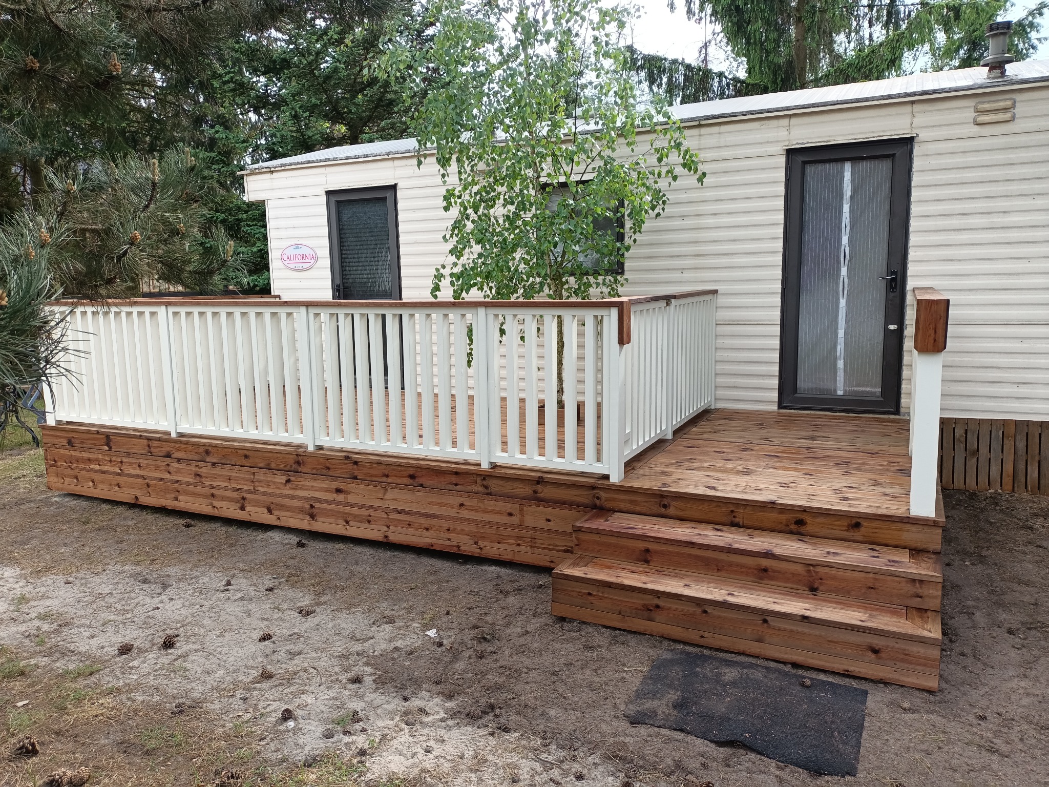 Drewniany taras z białą balustradą przy domu mobilnym. Widoczne schody wejściowe i drzewo zasadzone obok tarasu. Całość w naturalnej kolorystyce drewna i bieli.