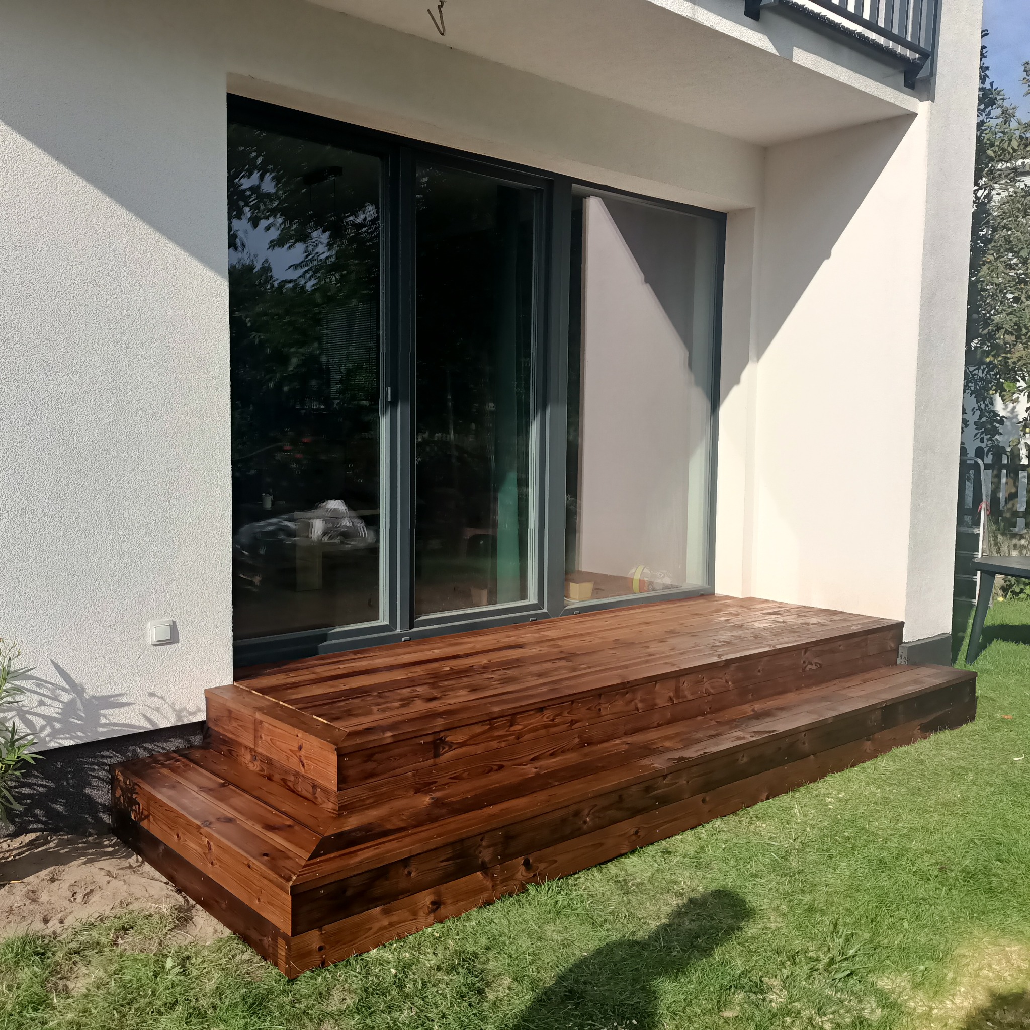 Nowoczesny taras drewniany z ciemnego drewna, prowadzący do przeszklonych drzwi domu. Widoczne schody tarasowe i trawnik. Jasne oświetlenie podkreśla strukturę drewna.