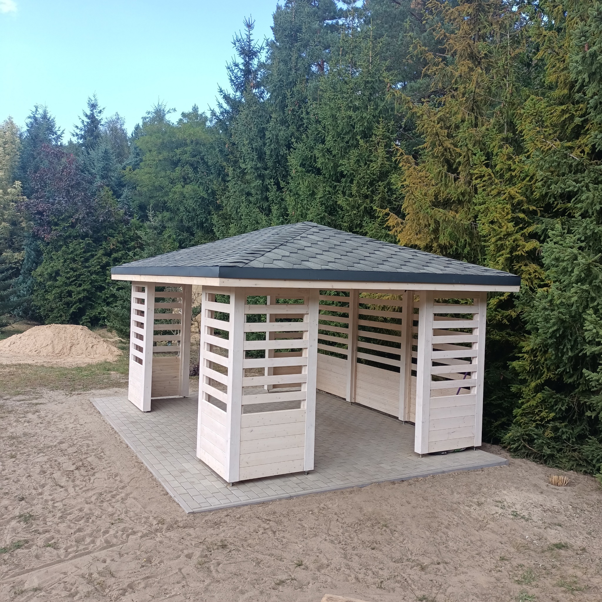 Nowoczesna, jasna altana z szarym dachem na tle zielonych drzew. Konstrukcja z drewnianymi panelami i kostką brukową. Widoczny piasek i elementy otoczenia.