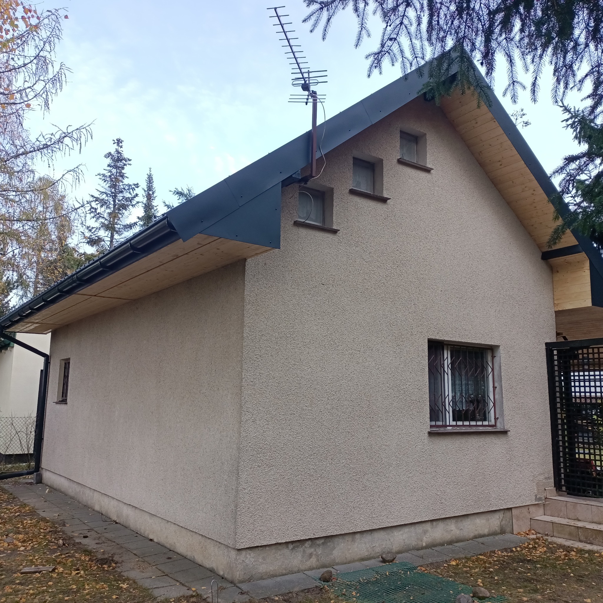 Wykończony budynek z elewacją w odcieniach beżu, z ciemnym dachem i anteną. Widoczne okna z kratami i fragment drzewa. Ujęcie z boku budynku.