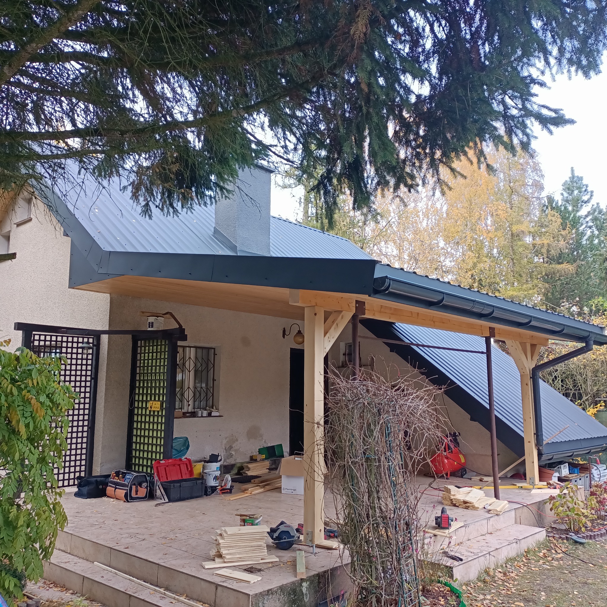 W trakcie budowy zadaszenia tarasu przy domu. Widoczne drewniane słupy, fragment dachu z blachy, narzędzia i materiały budowlane na tarasie. Jesienne otoczenie.