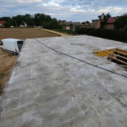 OptimusDom - Wylana płyta fundamentowa z widocznym szalunkiem i wystającymi elementami konstrukcyjnymi, obok zaparkowany biały bus dostawczy, w tle zabudowa jednorodzinna i pochmurne niebo.