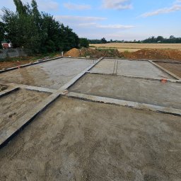 OptimusDom - Przygotowany zbrojony fundament pod budowę domu jednorodzinnego, widoczne szalunki wypełnione piaskiem i gruzem, w tle drzewa i pole uprawne pod błękitnym niebem.