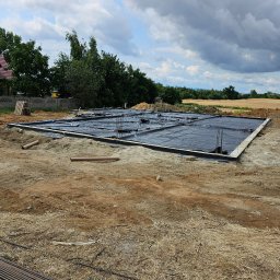 OptimusDom - Przygotowany teren pod wylewkę fundamentów: widoczne deskowanie, izolacja w postaci czarnej folii oraz wystające pręty zbrojeniowe na tle pola i pochmurnego nieba.