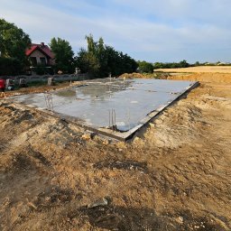 OptimusDom - Wylany fundament płyty betonowej z wystającymi prętami zbrojeniowymi, otoczony wykopem ziemnym, w tle dom jednorodzinny i pole uprawne pod błękitnym niebem.