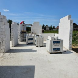 OptimusDom - Wznoszenie ścian budynku z bloczków silikatowych SIL-PRO, widoczny pracownik w czerwonej koszulce oraz palety z bloczkami na placu budowy w słoneczny dzień.