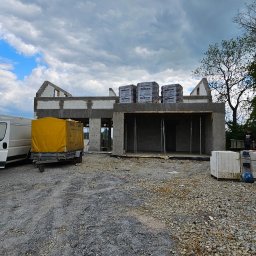 OptimusDom - Surowy stan domu w trakcie budowy z pustaków, widoczne bloczki Röben na dachu, przyczepka z żółtą plandeką, gruz na placu budowy i pochmurne niebo.