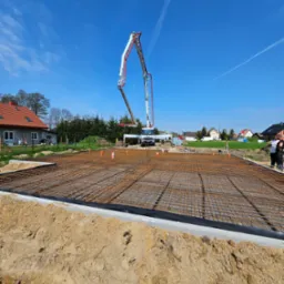 Zbrojenie fundamentów przygotowane do zalania betonem, widoczna pompa do betonu na tle błękitnego nieba, teren budowy w słoneczny dzień.