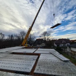 Żółty dźwig podnosi element stropu nad budowanym domem; widoczne prefabrykowane płyty stropowe z wystającymi prętami zbrojeniowymi, ceglane wypełnienia i drabina; w tle zabudowa jednorodzinna...