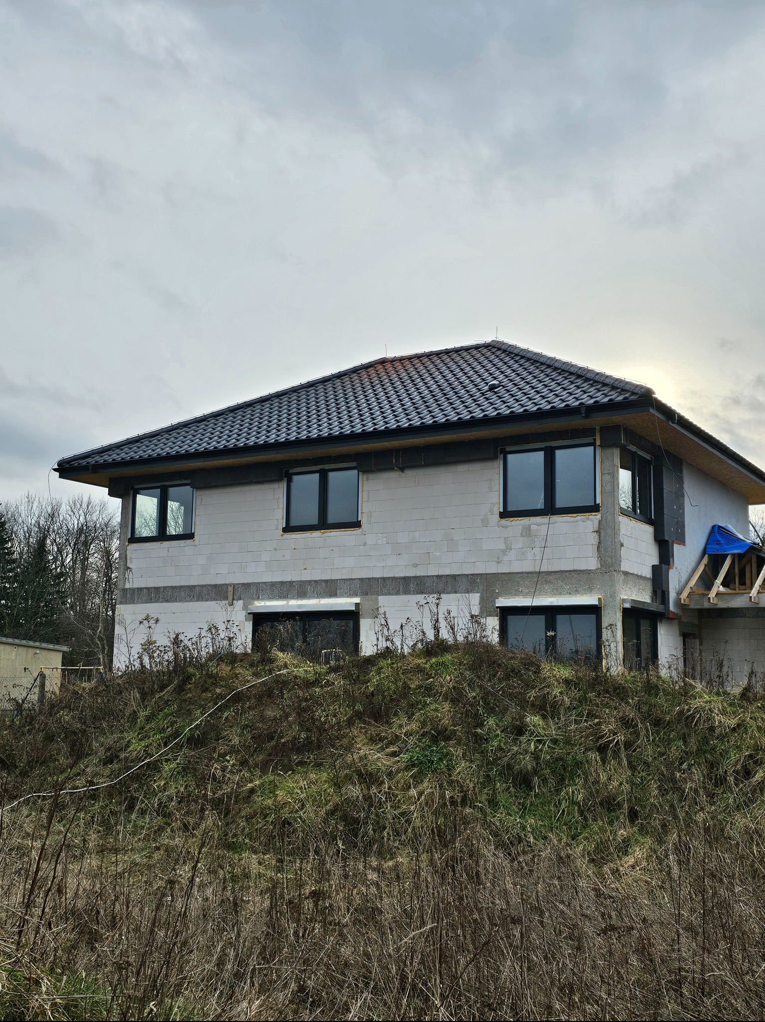 Surowy stan domu z białych bloczków, czarne okna, dachówka ceramiczna, widoczne elementy konstrukcyjne dachu, otoczenie zarośnięte trawą i chwastami, pochmurne niebo.