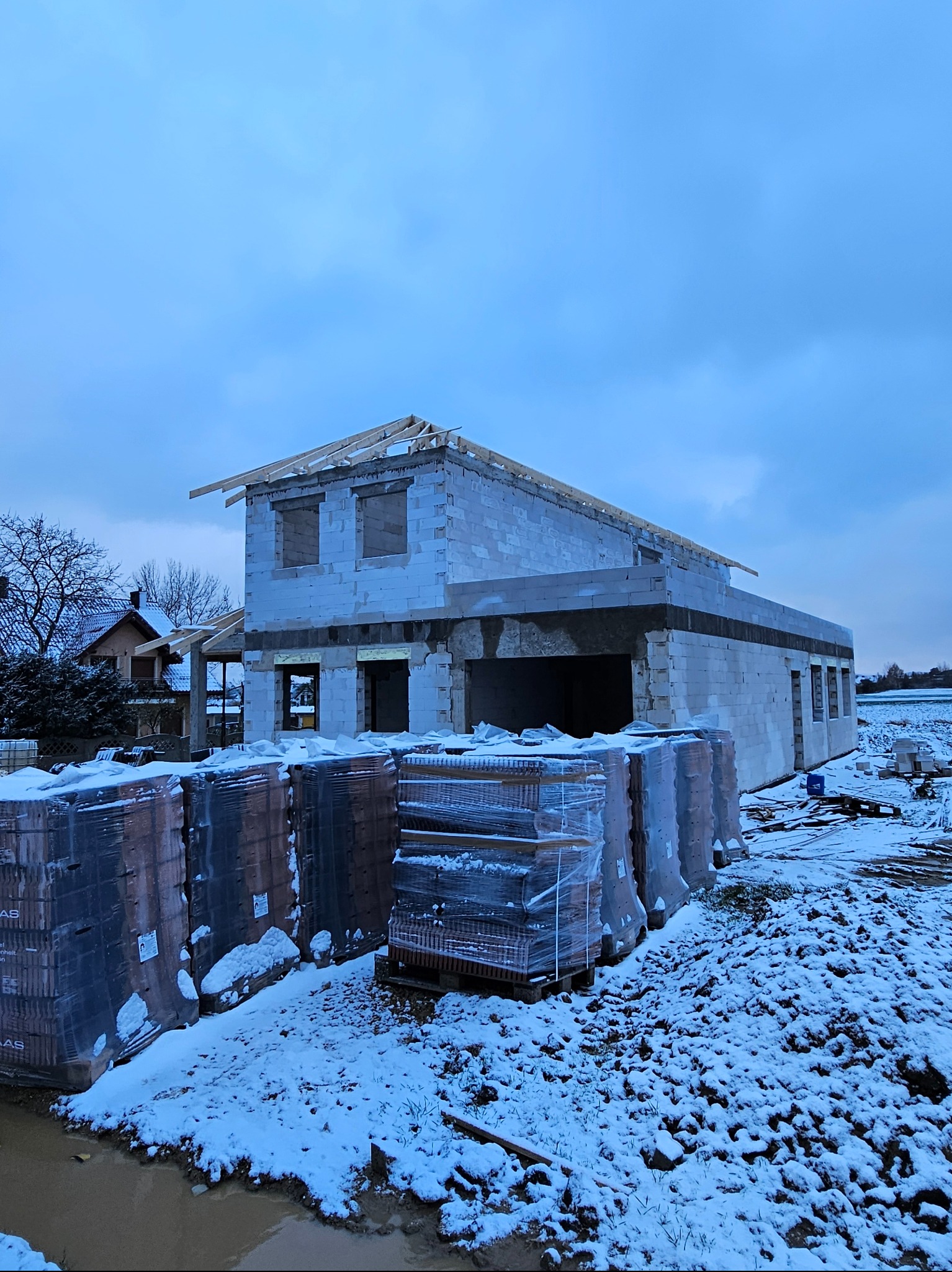 Niedokończony dwukondygnacyjny budynek z pustaków w zimowej scenerii, z widoczną konstrukcją dachu i paletami cegieł w folii na pierwszym planie.