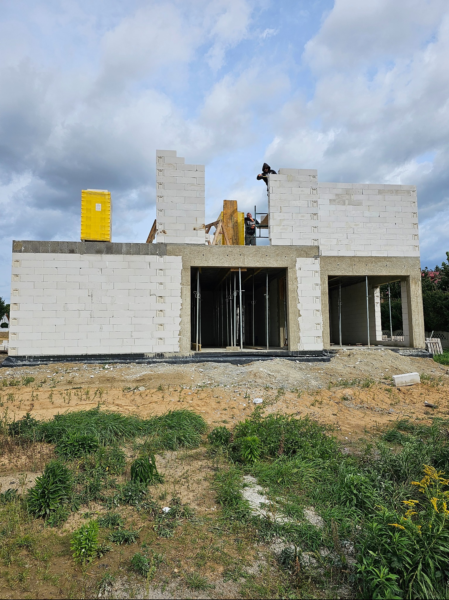 Wznoszenie ścian dwukondygnacyjnego domu z białych bloczków, widoczni dwaj pracownicy na rusztowaniu, żółta paleta z materiałami na stropie, teren wokół w stanie surowym.