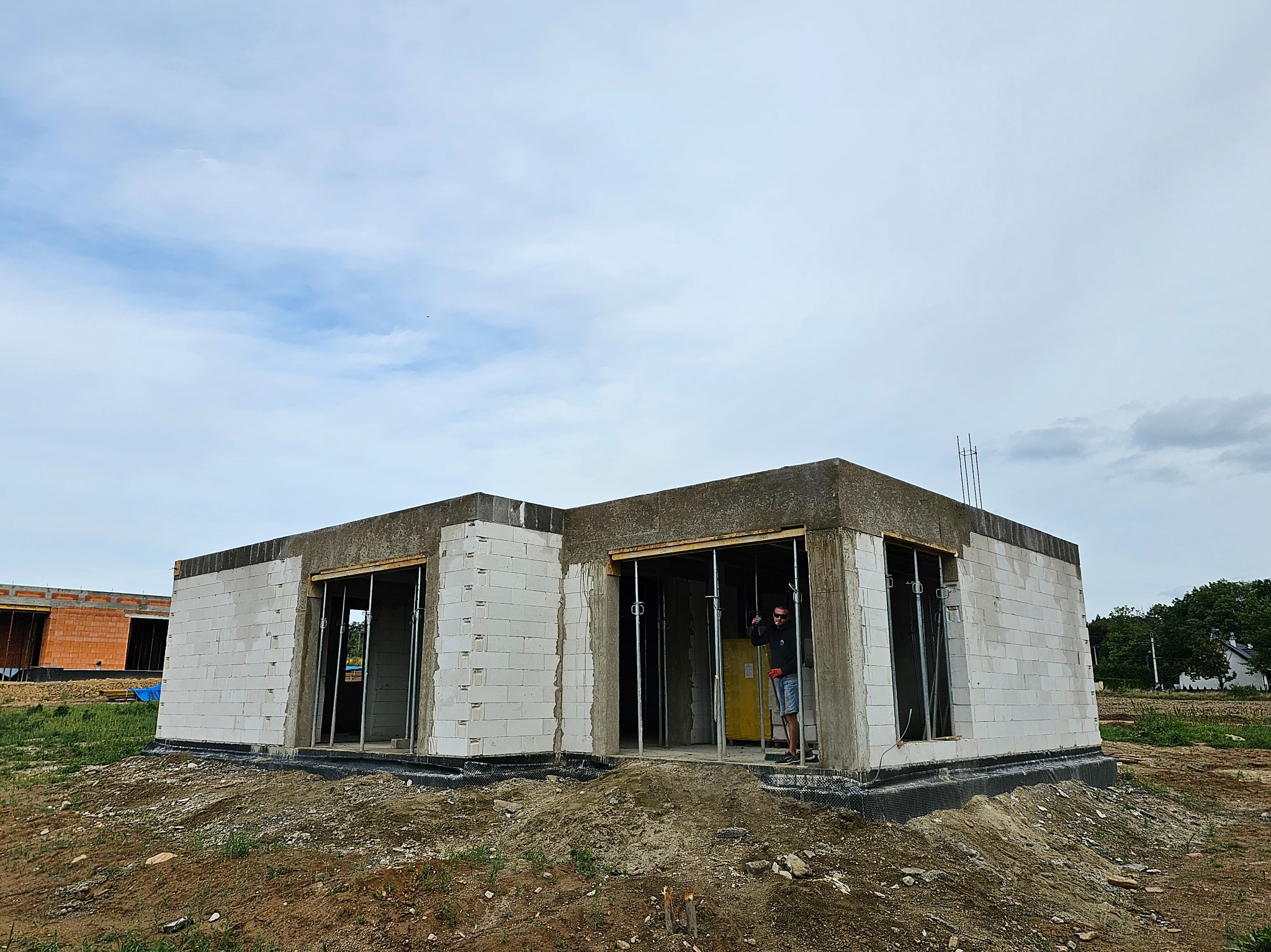 Niski, parterowy budynek w stanie surowym otwartym, wzniesiony z białych bloczków, widoczny pracownik w wejściu, teren wokół budynku nierówny, w tle częściowo widoczny inny budynek w trakcie budowy.