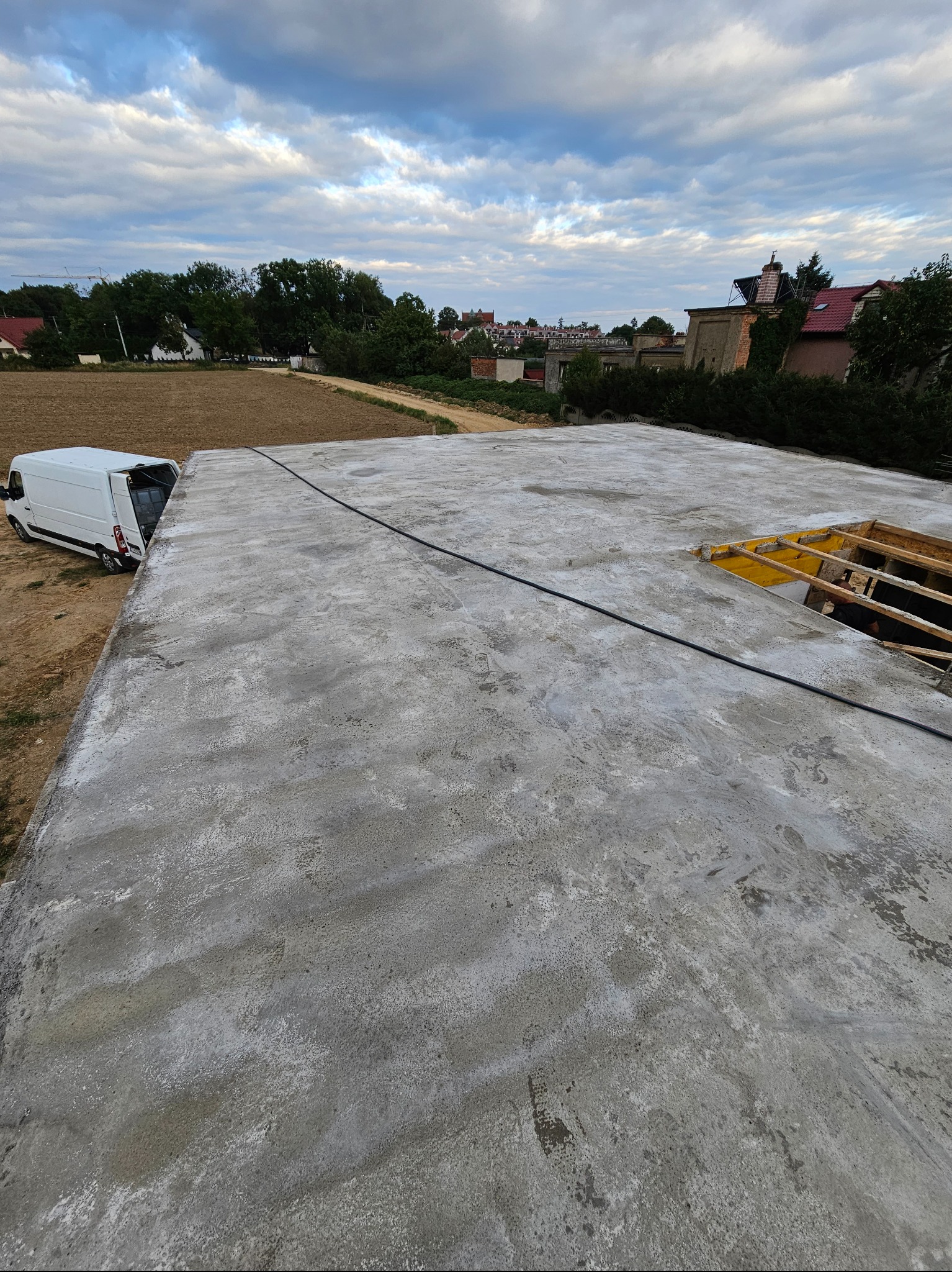 Wylana płyta fundamentowa z widocznym szalunkiem i wystającymi elementami konstrukcyjnymi, obok zaparkowany biały bus dostawczy, w tle zabudowa jednorodzinna i pochmurne niebo.