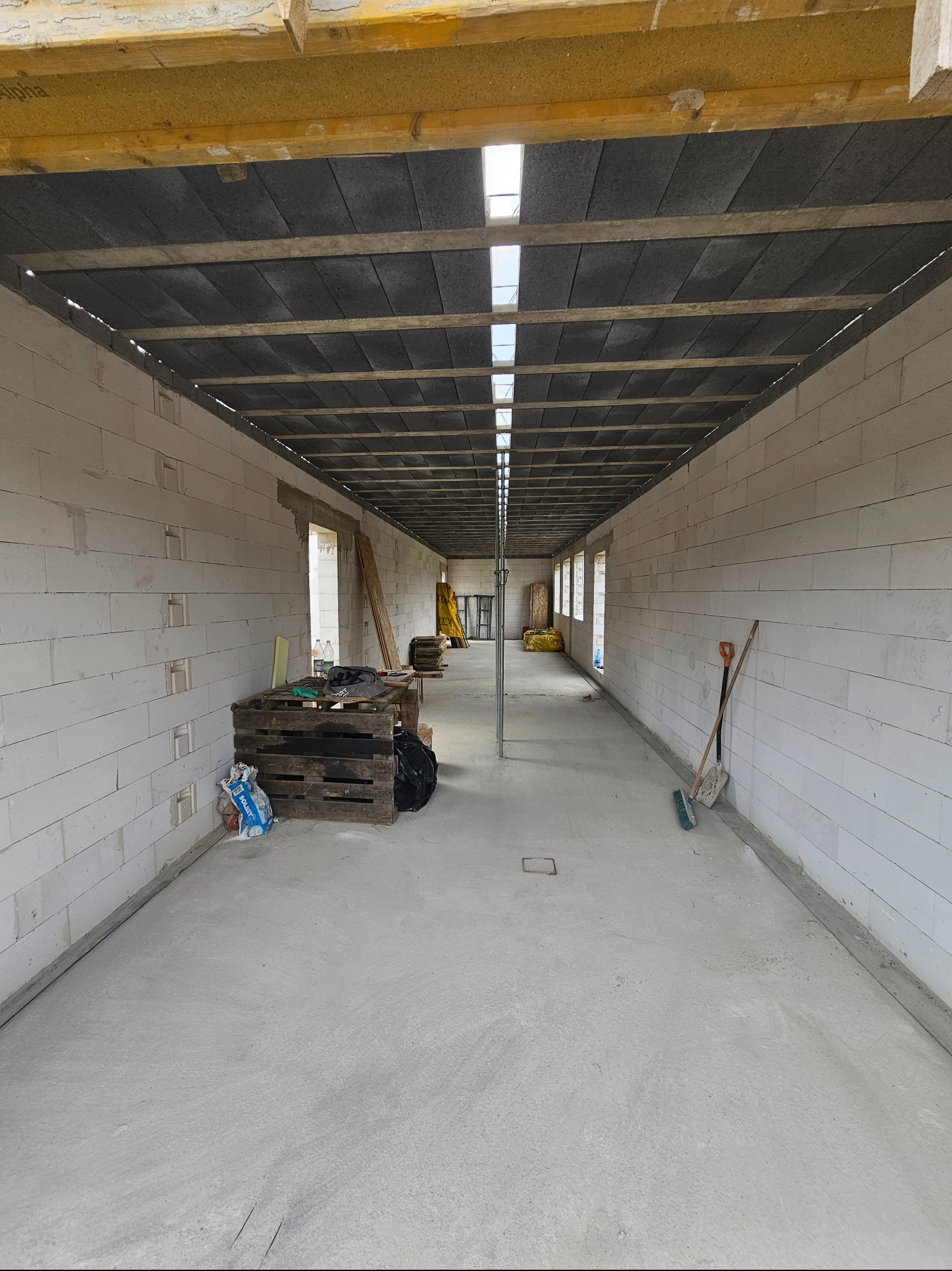 Wnętrze domu w trakcie budowy, ściany z białych bloczków, betonowa posadzka, strop z drewnianych belek i izolacji, widoczne narzędzia i materiały budowlane.