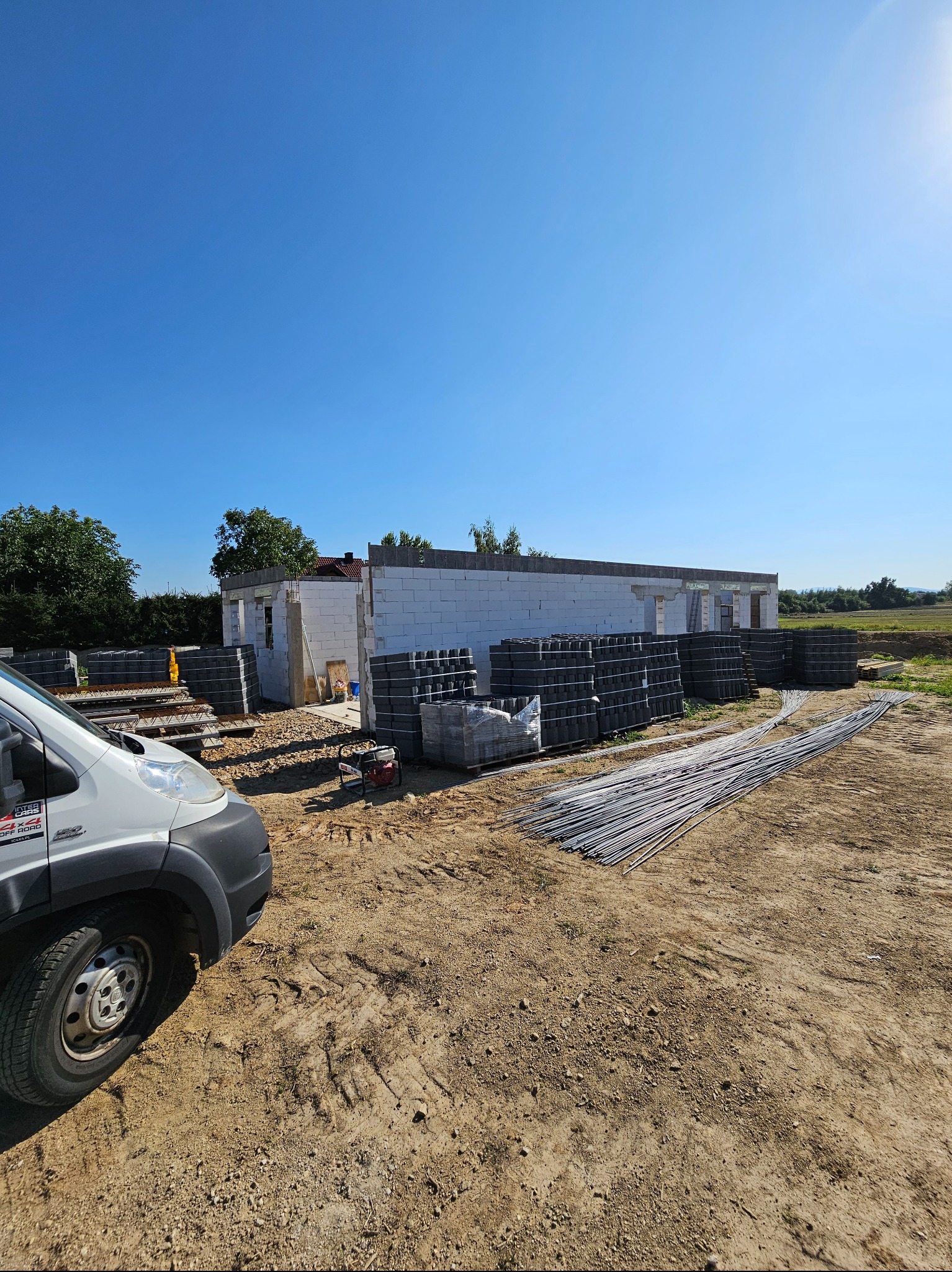 Wznoszenie ścian parterowego domu z bloczków betonu komórkowego, widoczne zbrojenia i palety z materiałem budowlanym, na pierwszym planie samochód dostawczy.