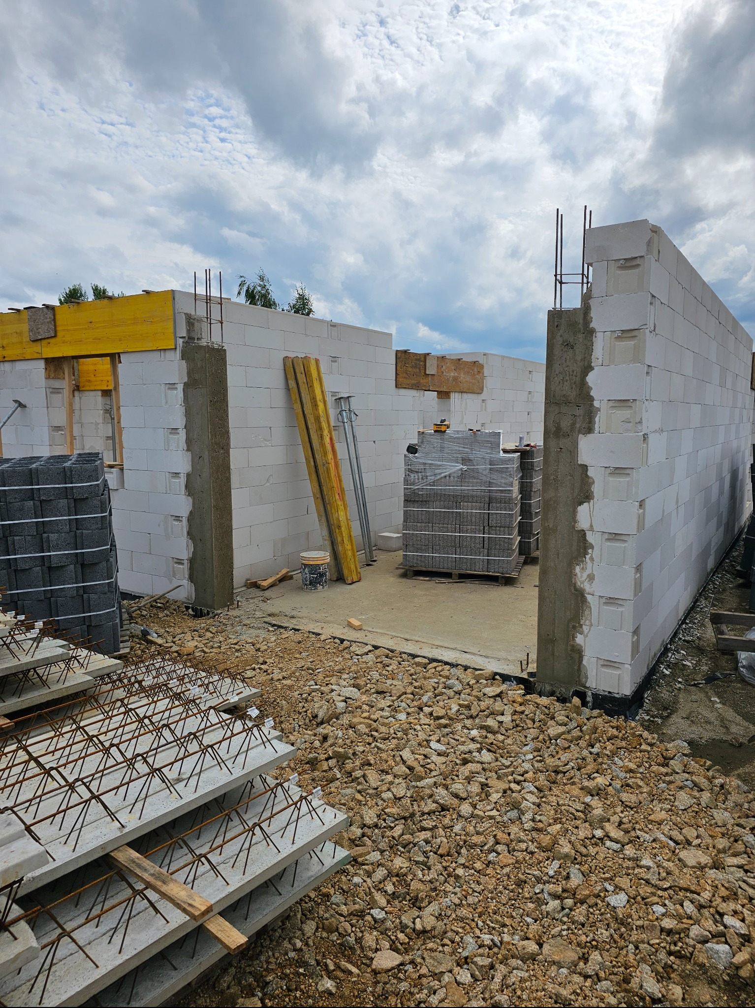 Wznoszenie ścian parterowego domu z bloczków betonu komórkowego, widoczne zbrojenia pionowe, szalunki stropowe i materiały budowlane na placu budowy.