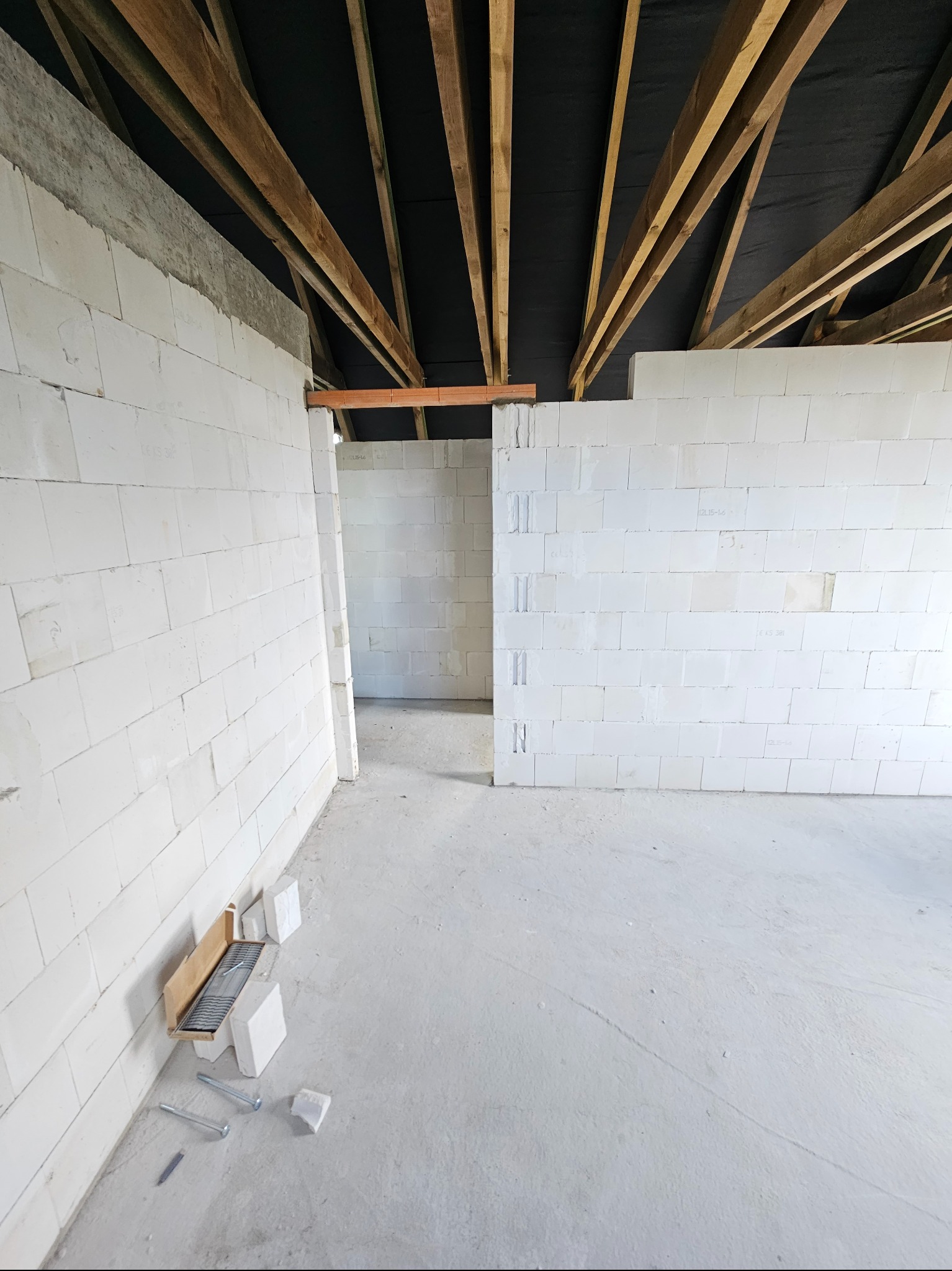 Wnętrze domu w trakcie budowy z białych bloczków, widoczne drewniane elementy konstrukcji dachu i narzędzia murarskie na podłodze.