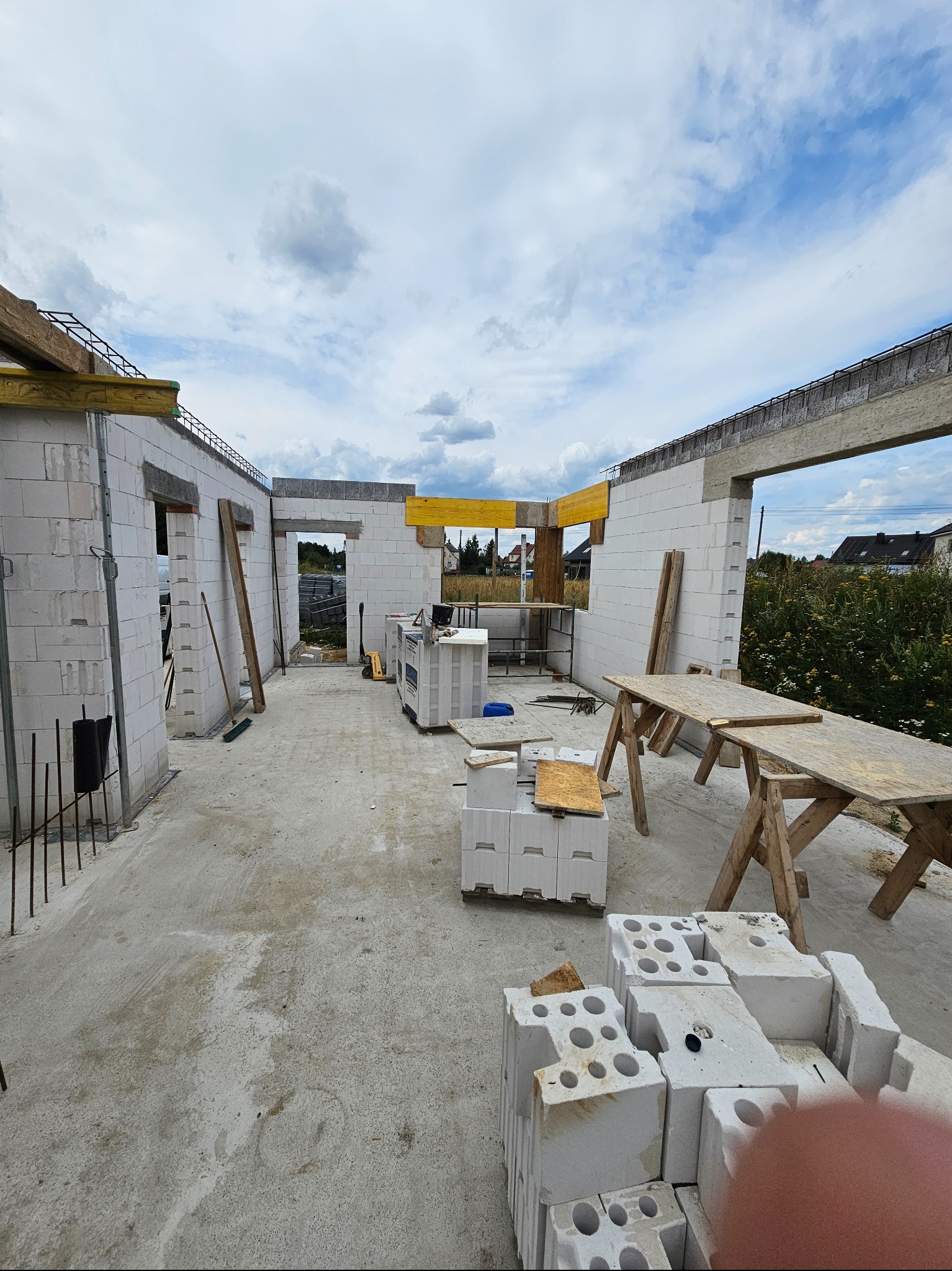 Wnętrze surowego, murowanego domu w trakcie budowy. Widoczne ściany z betonu komórkowego, drewniane stemple i stropy, a także stosy bloczków. W oddali widać teren działki i zabudowania.