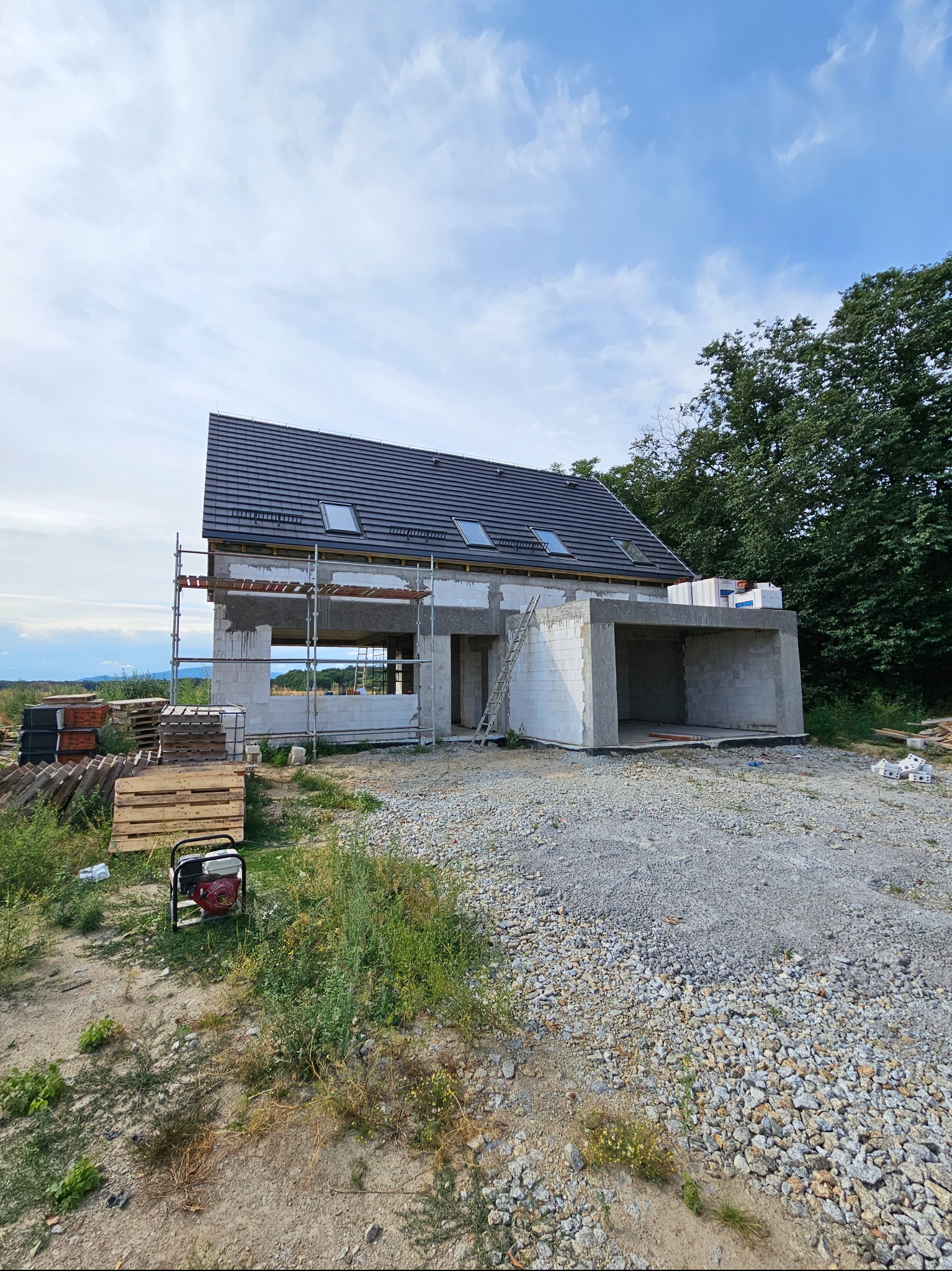 Surowy stan domu jednorodzinnego z szarej cegły, z widocznym rusztowaniem, ułożonym dachem z oknami dachowymi, z generatorem prądu obok palet i gruzu na pierwszym planie.