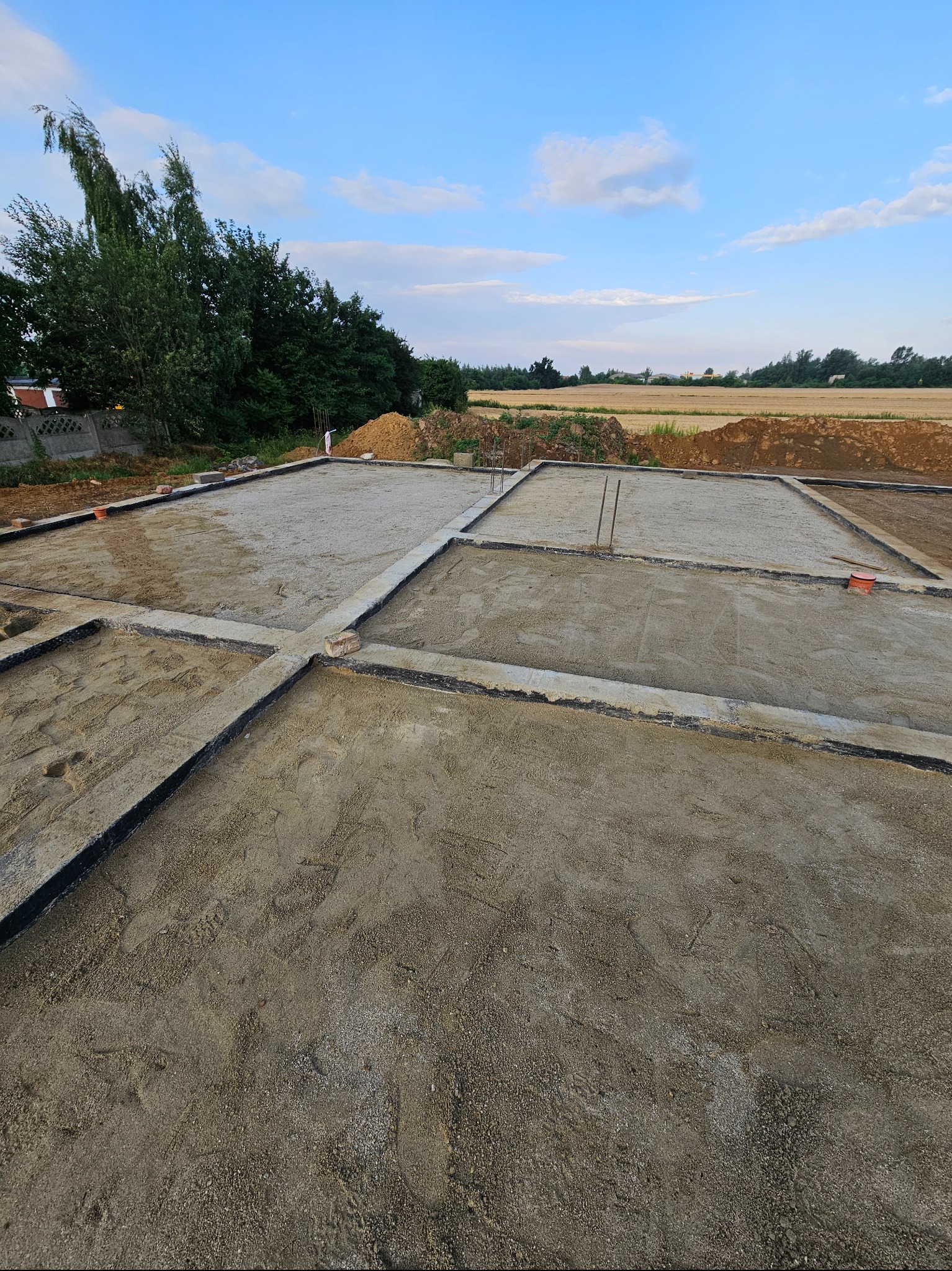 Przygotowany zbrojony fundament pod budowę domu jednorodzinnego, widoczne szalunki wypełnione piaskiem i gruzem, w tle drzewa i pole uprawne pod błękitnym niebem.