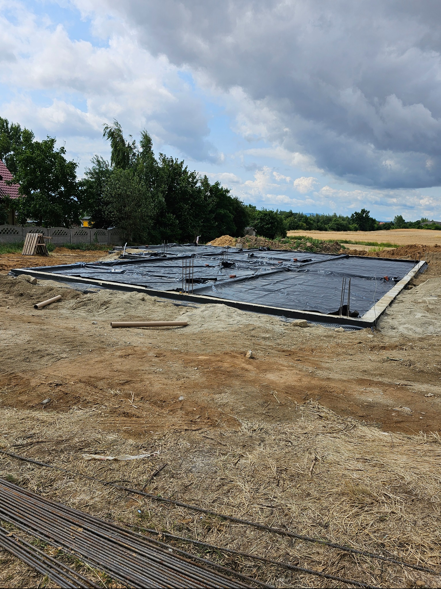 Przygotowany teren pod wylewkę fundamentów: widoczne deskowanie, izolacja w postaci czarnej folii oraz wystające pręty zbrojeniowe na tle pola i pochmurnego nieba.