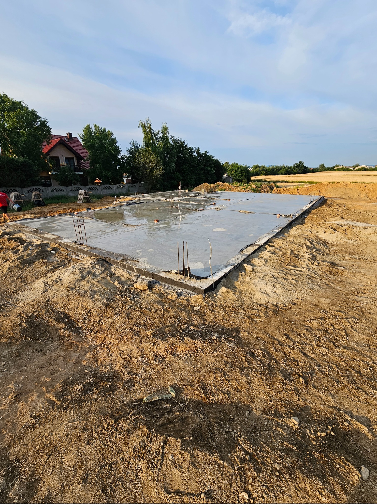 Wylany fundament płyty betonowej z wystającymi prętami zbrojeniowymi, otoczony wykopem ziemnym, w tle dom jednorodzinny i pole uprawne pod błękitnym niebem.