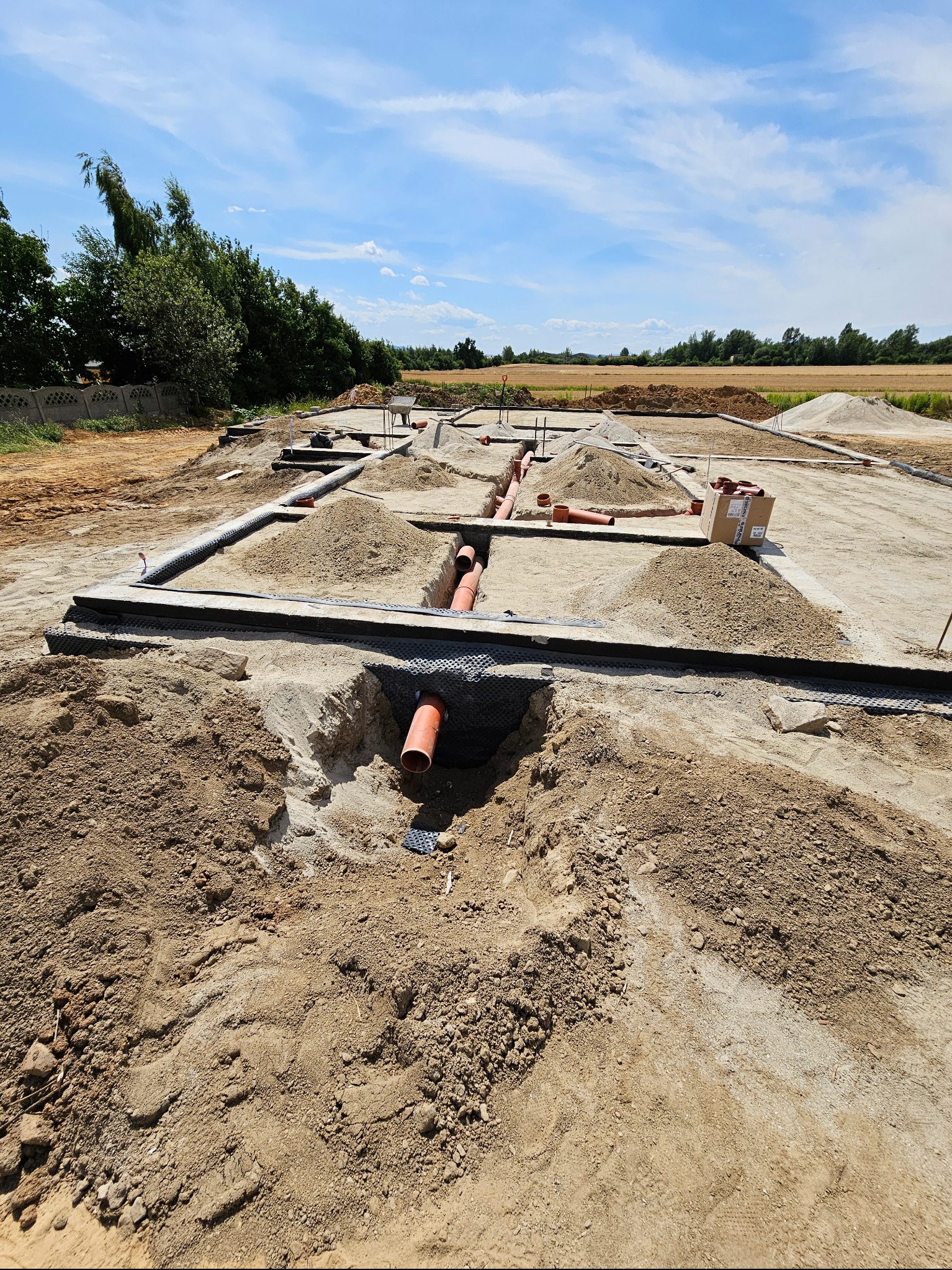 Przygotowanie terenu pod budowę domu: wykopane fundamenty z widocznymi rurami kanalizacyjnymi, zabezpieczone czarną folią, piasek zasypany wokół, w tle krajobraz polny pod błękitnym niebem.