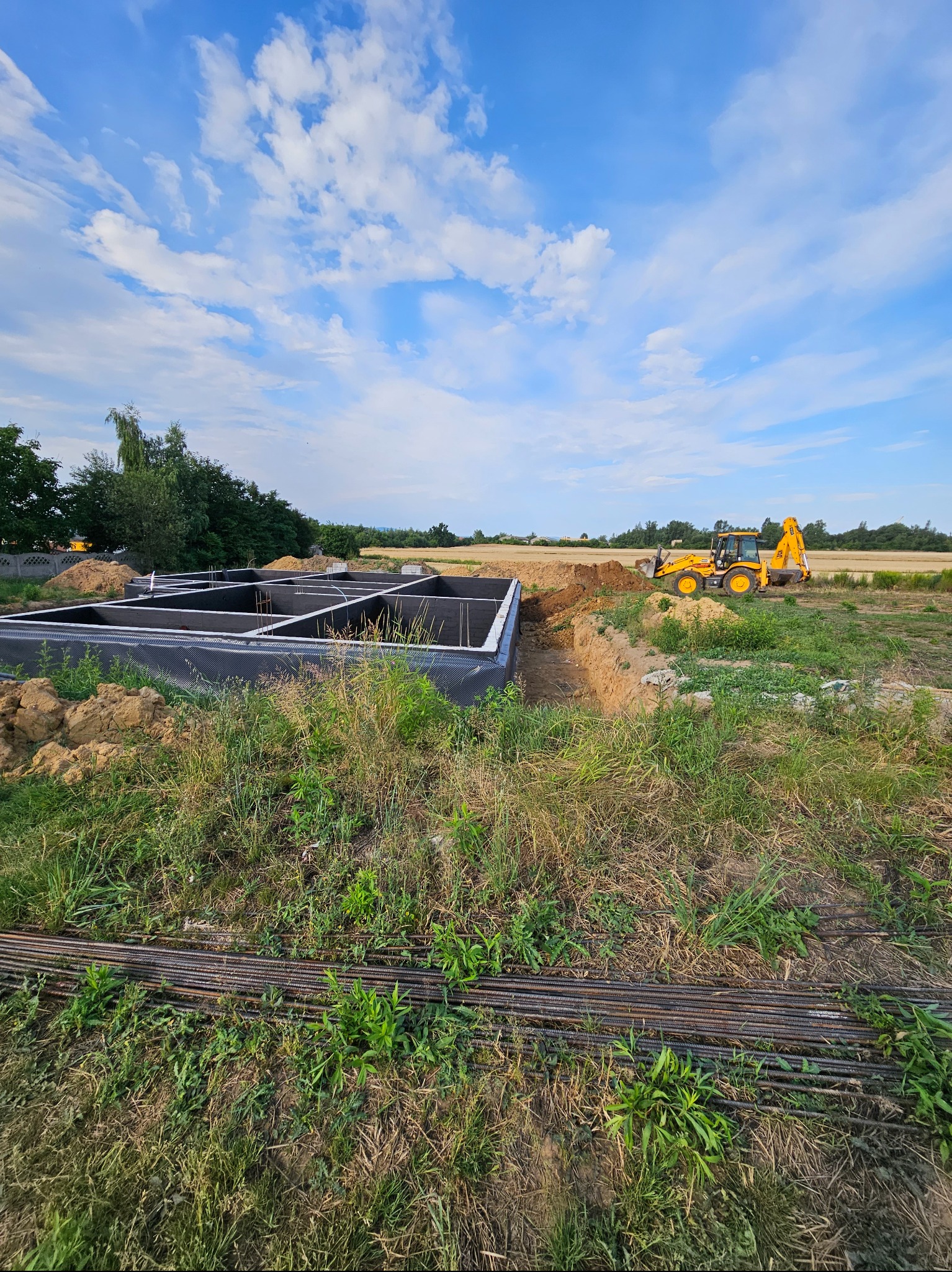 Przygotowany i zaizolowany fundament pod budowę domu, z wykopem wykonanym przez koparkę, widok z poziomu gruntu na tle błękitnego nieba z chmurami.