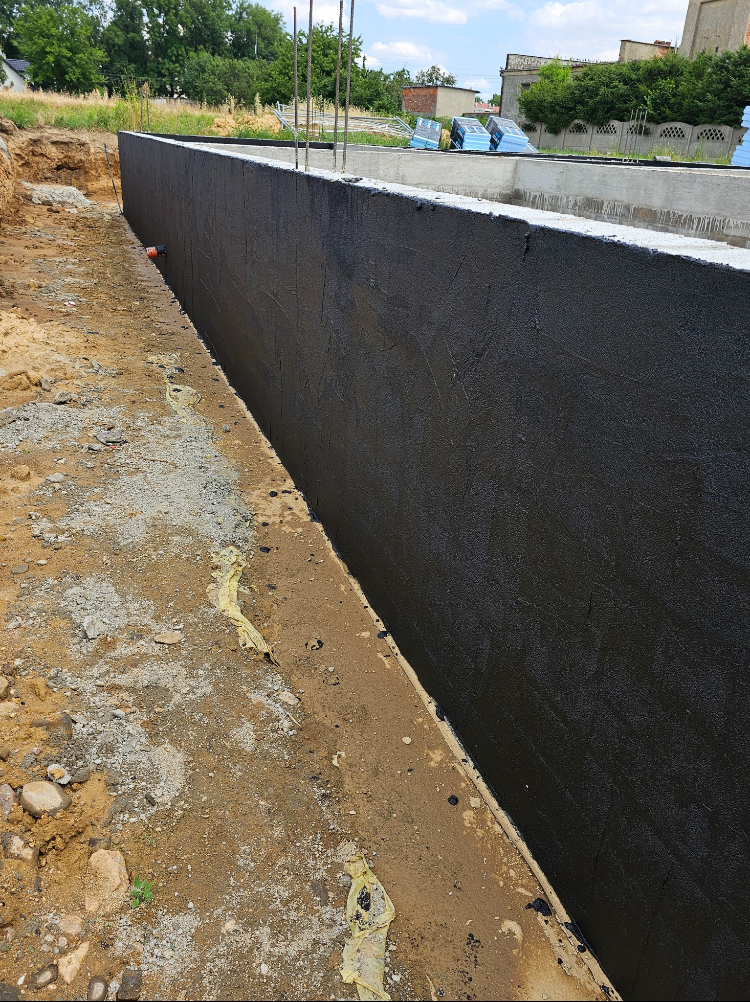 Świeżo zaizolowana, czarna, pionowa ściana fundamentowa z wystającymi prętami zbrojeniowymi, widoczna z bliska na tle wykopu i zieleni.