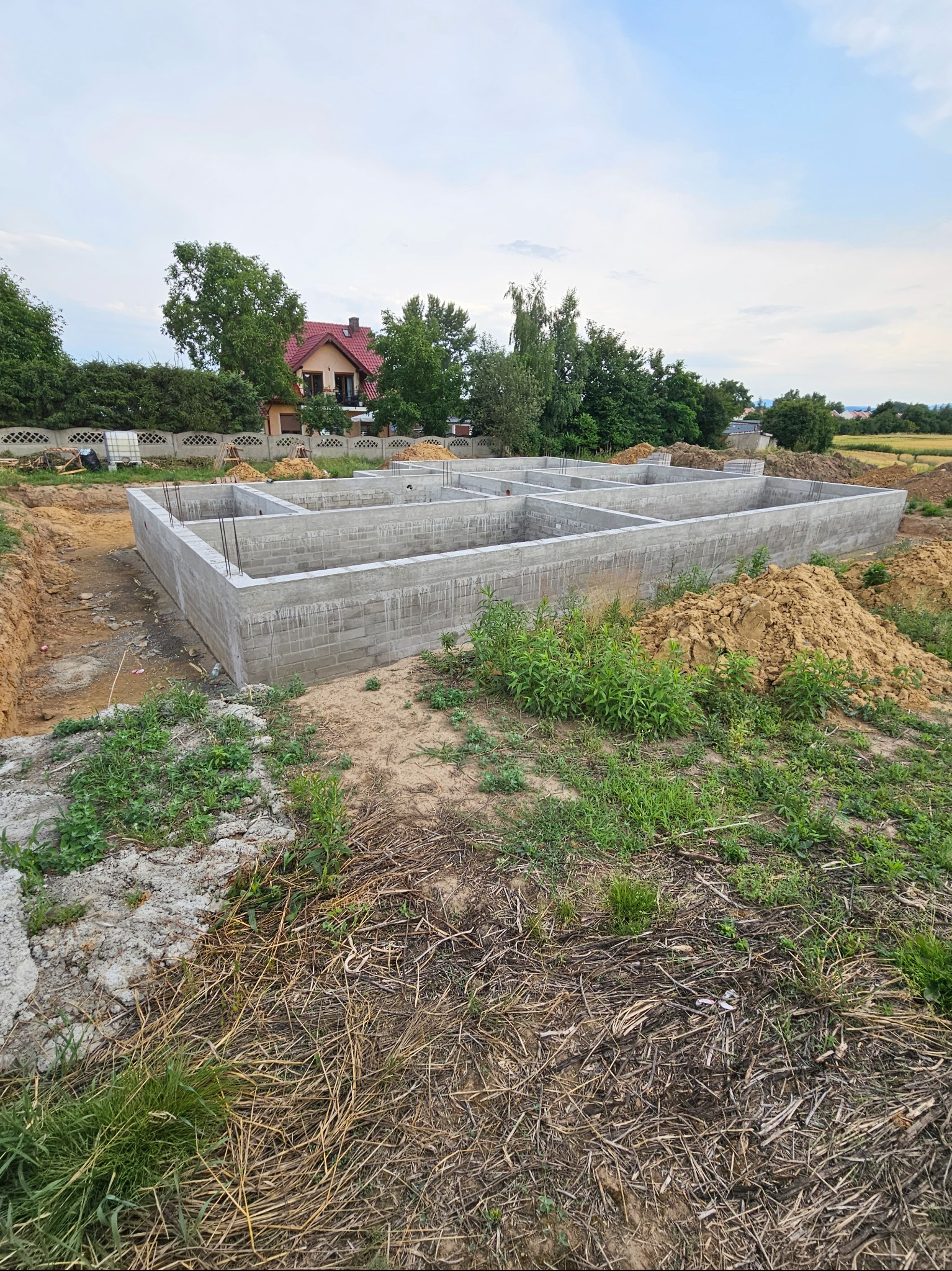 Surowy fundament z betonu komórkowego zbrojony stalą, widok z poziomu gruntu na tle zieleni i domu z czerwoną dachówką.
