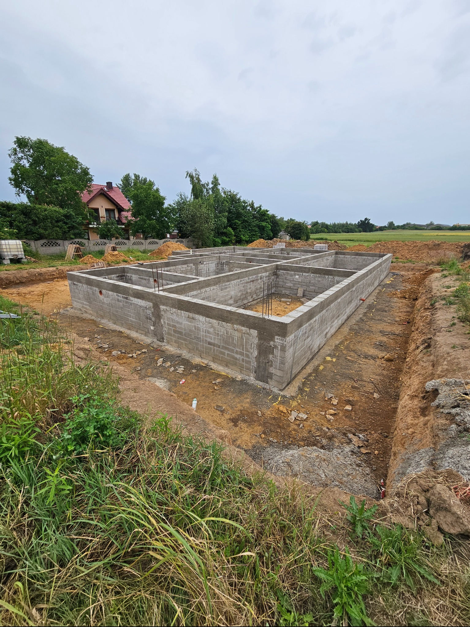 Surowy, betonowy fundament domu z widocznymi prętami zbrojeniowymi, otoczony wykopem i zielenią, na tle domu z czerwoną dachówką pod pochmurnym niebem.