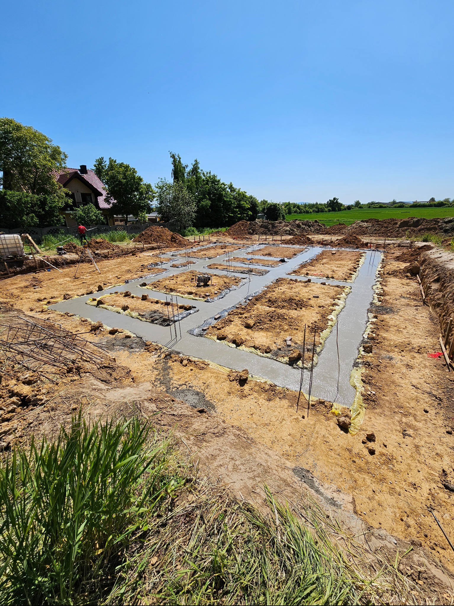 Wylane betonowe ławy fundamentowe z wystającymi prętami zbrojeniowymi, zabezpieczone folią, na tle krajobrazu z domem i zielenią.