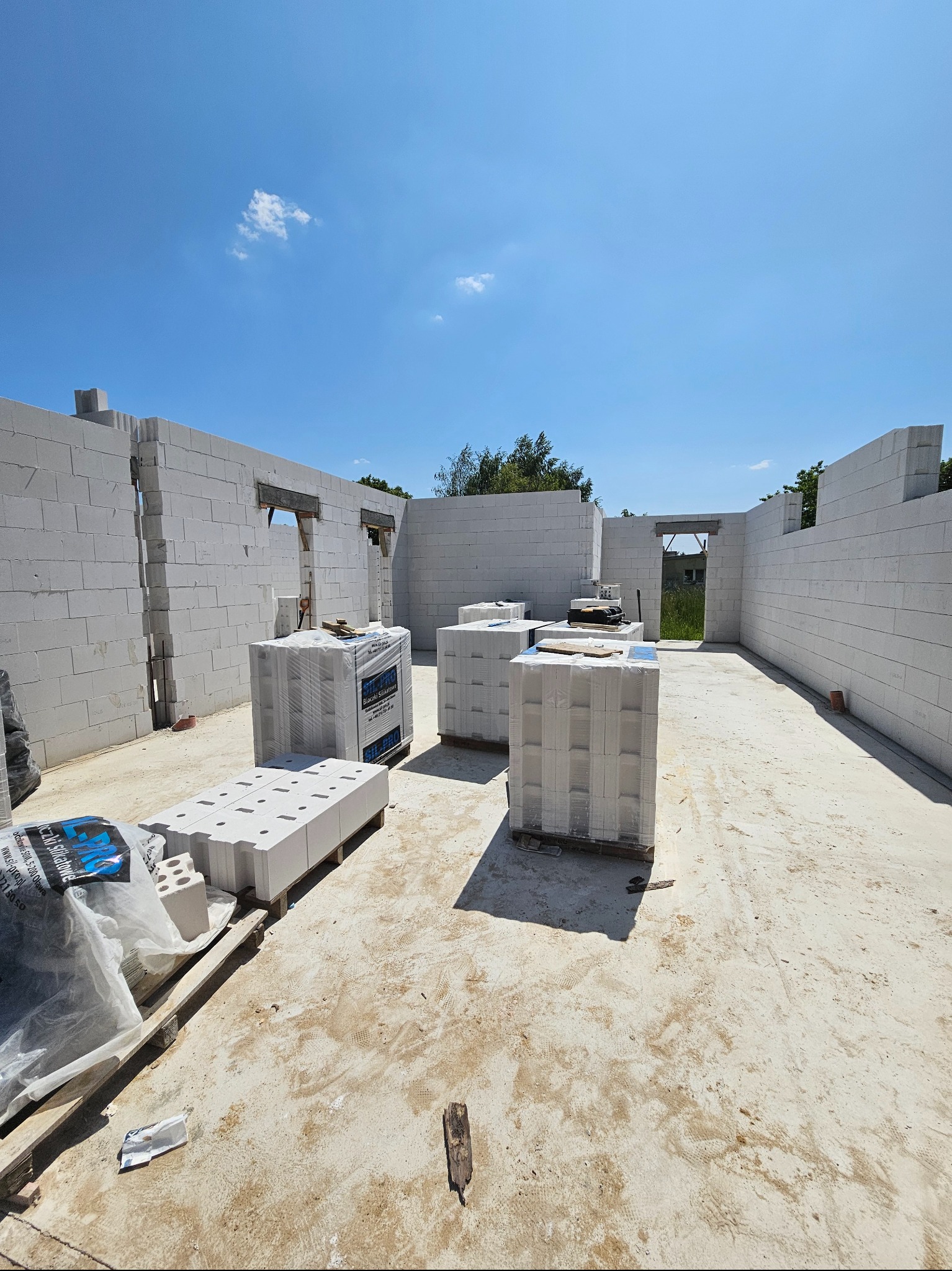 Wnętrze wznoszonego domu z białych bloków silikatowych, palety z zapakowanymi bloczkami na betonowej posadzce, widoczne otwory okienne i drzwiowe, błękitne niebo w tle.