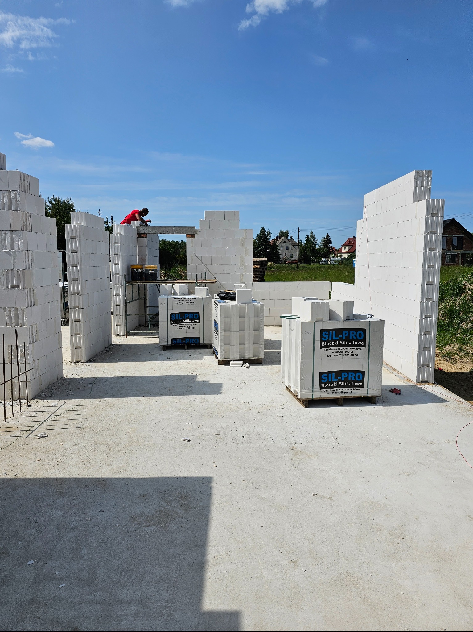 Wznoszenie ścian budynku z bloczków silikatowych SIL-PRO, widoczny pracownik w czerwonej koszulce oraz palety z bloczkami na placu budowy w słoneczny dzień.
