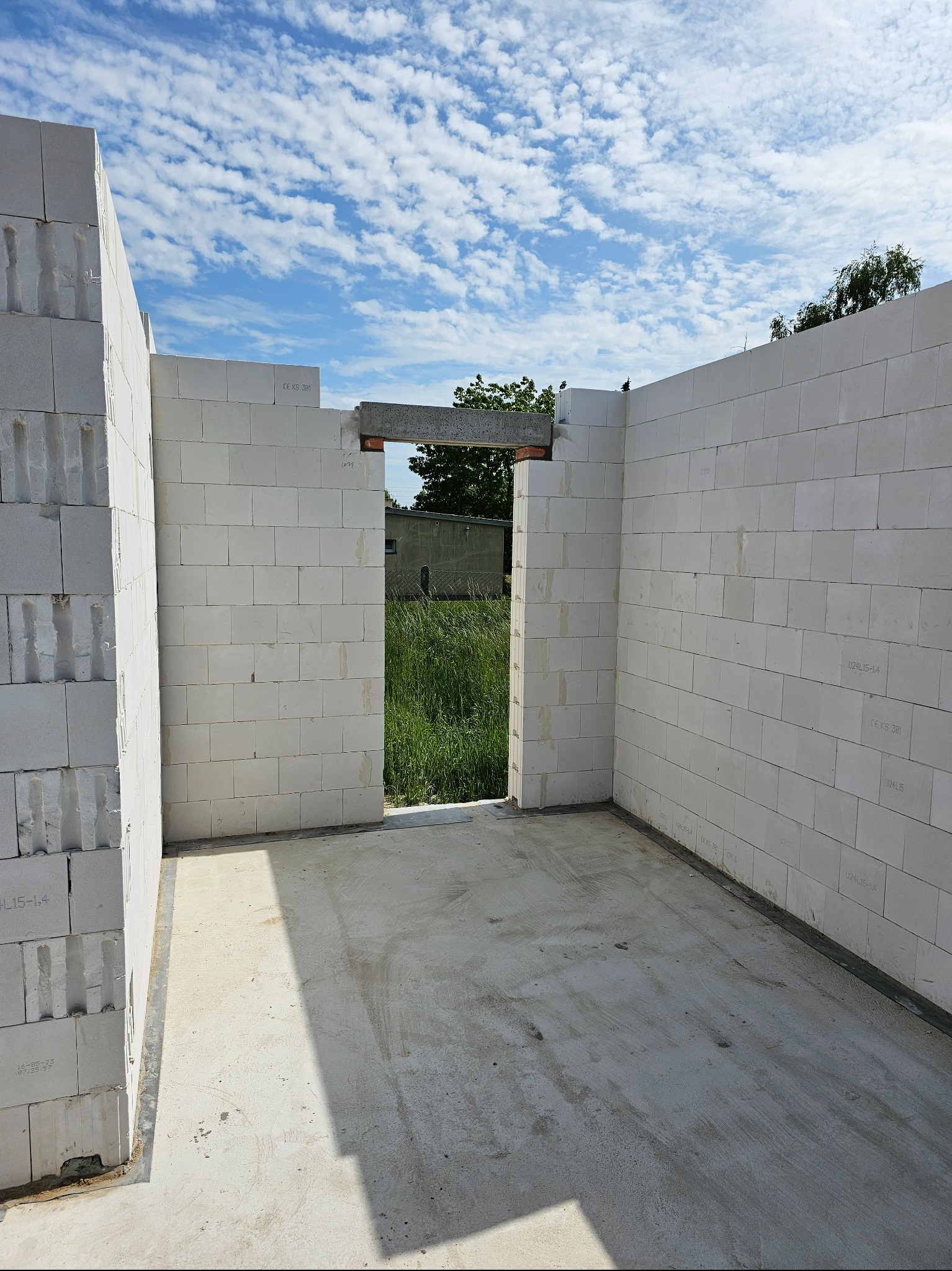 Wnętrze wznoszonego domu z białych bloczków, widoczne fragmenty ścian z oznaczeniami producenta, surowy betonowy fundament i fragment krajobrazu za otworem drzwiowym pod błękitnym niebem.