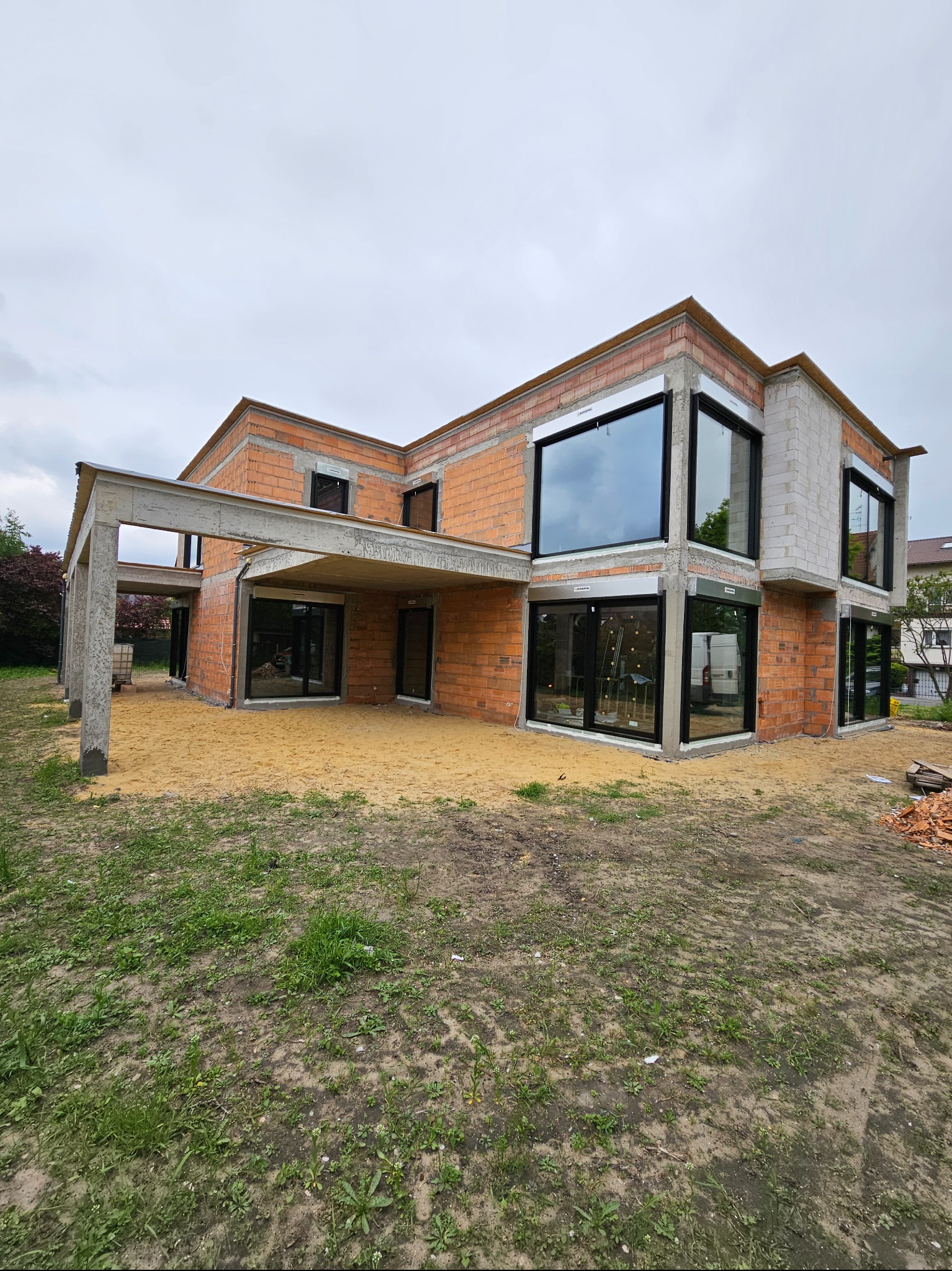 Surowy stan budynku z czerwonej cegły i białych bloczków, z dużymi oknami z czarnymi ramami, betonowym zadaszeniem i piaskowym podłożem, w tle widać samochód dostawczy.