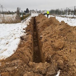 Stanisław Muzyka - Wąski wykop w ziemi z resztkami śniegu, w tle pracownik w żółtej kamizelce i biały samochód dostawczy na tle pochmurnego nieba i ogrodzenia.