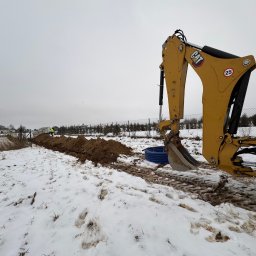 Stanisław Muzyka - Żółta koparka CAT przy wykopie w zasnieżonym terenie, widoczne niebieskie rury i pracownik w oddali.