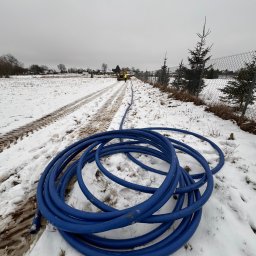 Stanisław Muzyka - Zwój niebieskiego węża na śniegu, w tle wykop i koparka w zimowej scenerii, widoczne ślady opon na ziemi.