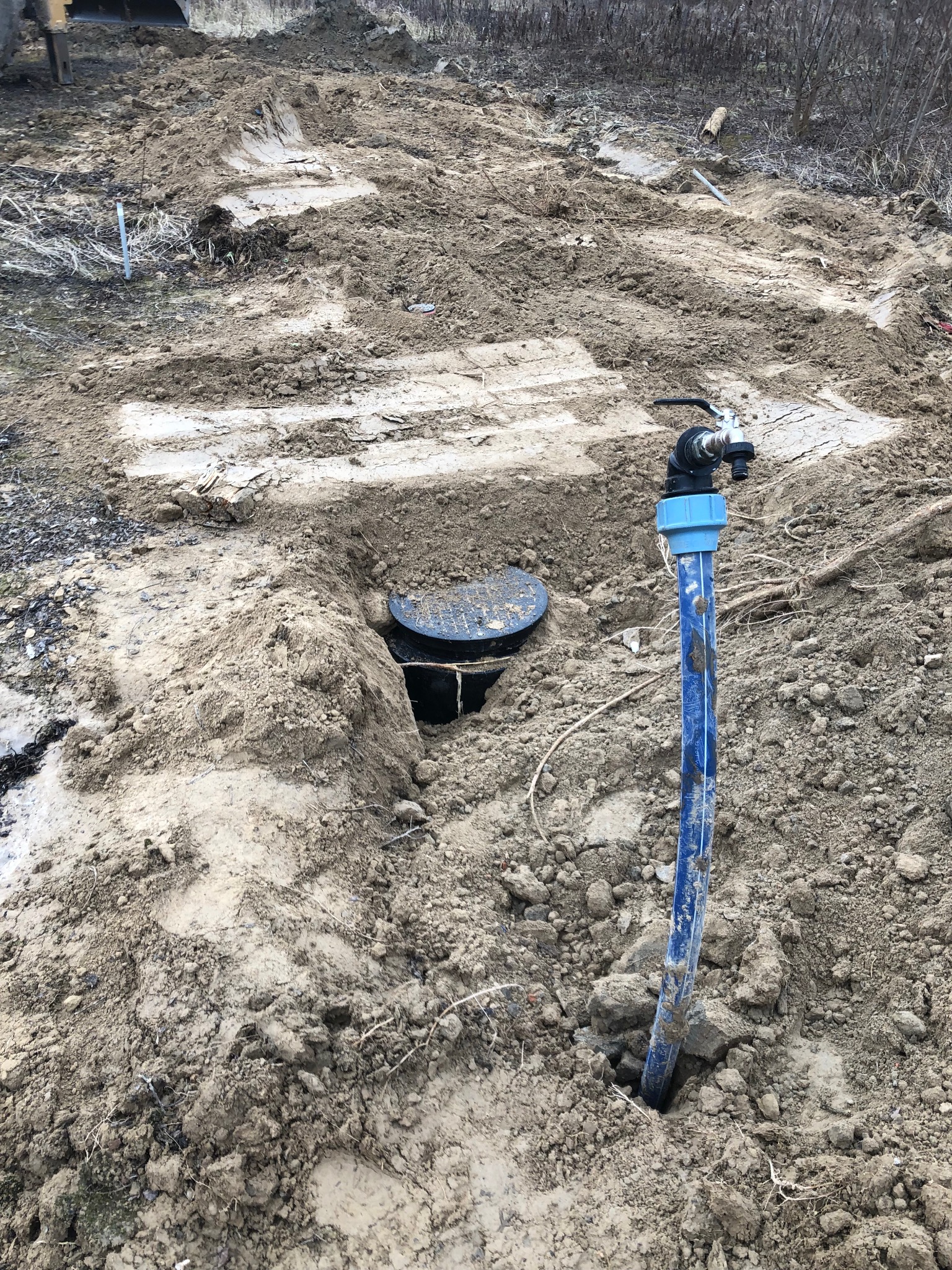 Niebieska rura z zaworem wystaje z ziemi obok włazu kanalizacyjnego na wykopanym terenie, widoczne ślady opon.