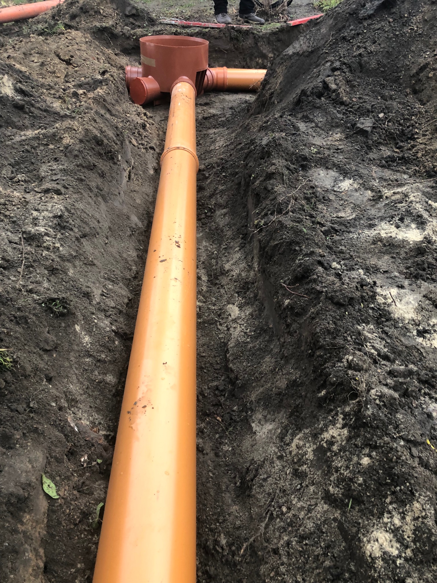 Pomarańczowa rura kanalizacyjna ułożona w wykopie ziemnym, widoczne elementy studzienki inspekcyjnej i przyłącza boczne.