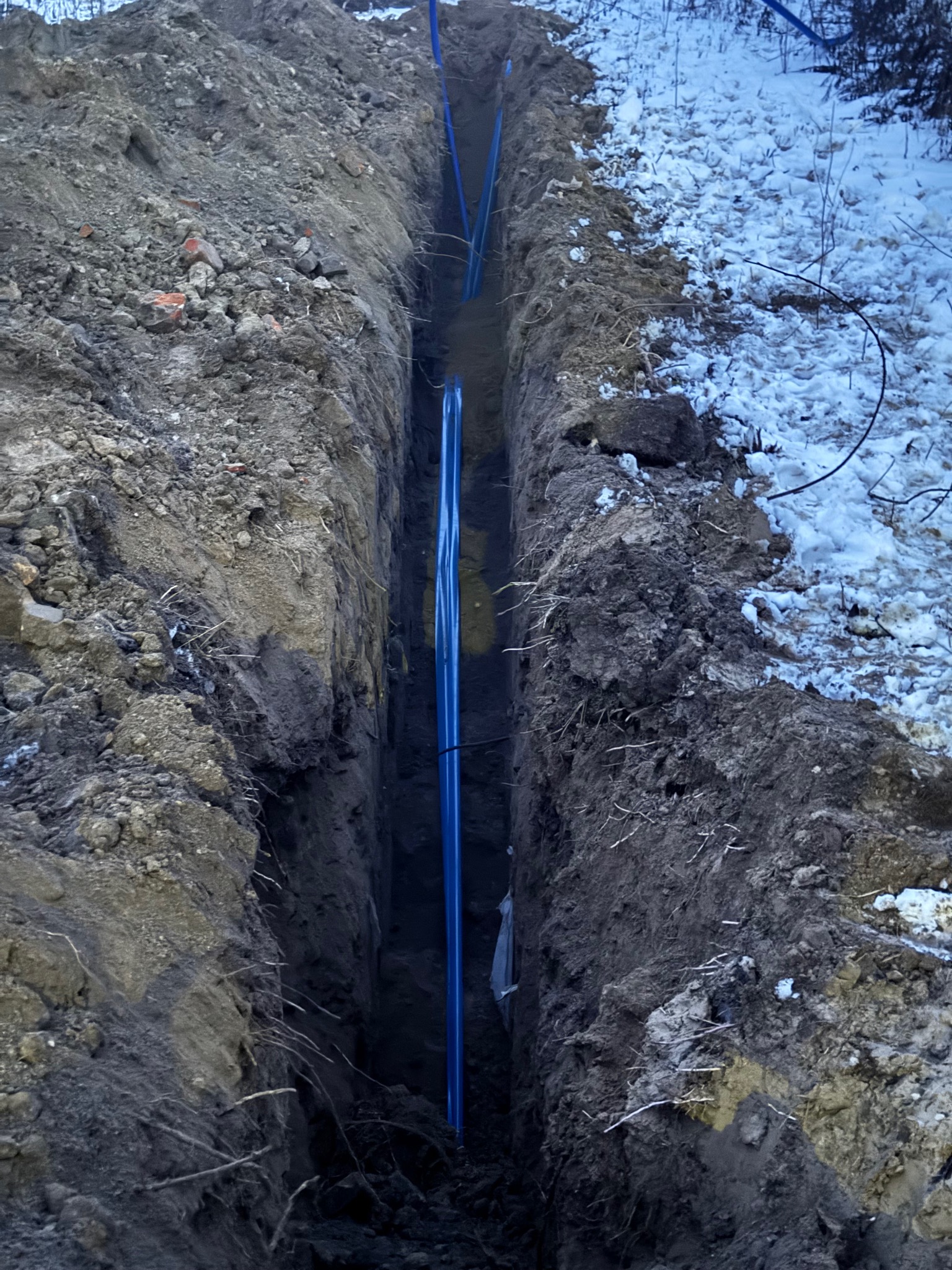 Głęboki wykop w ziemi z ułożonymi niebieskimi rurami, częściowo zasypany śniegiem po jednej stronie.