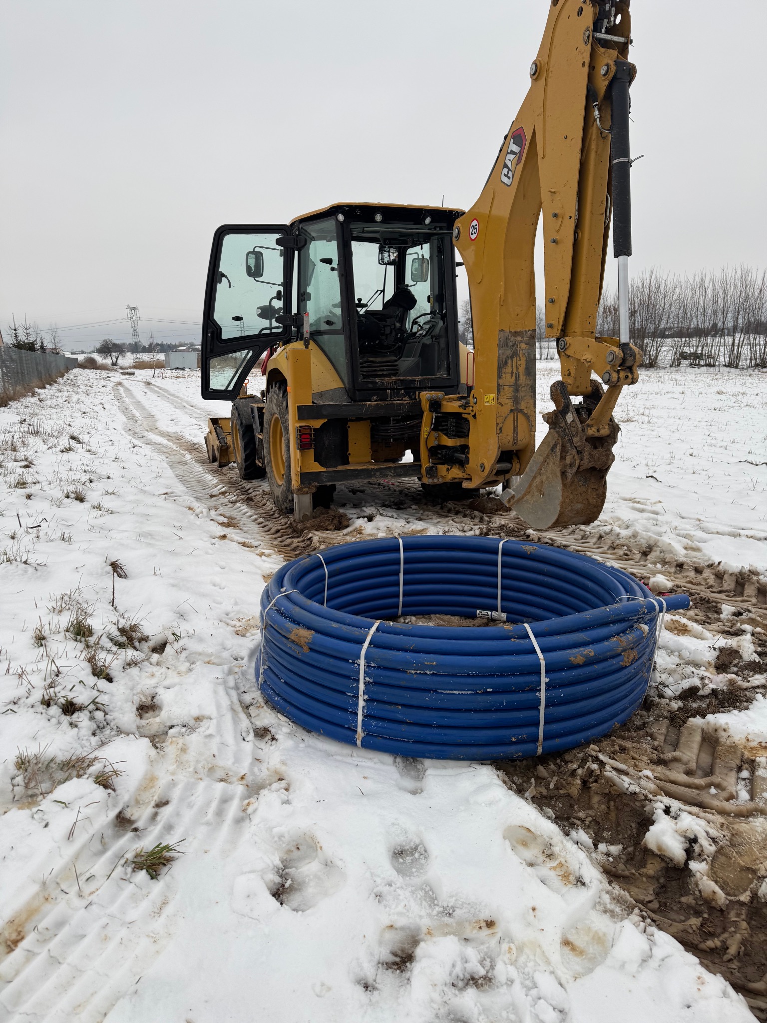 Żółta koparko-ładowarka CAT na zasnieżonym terenie z otwartymi drzwiami kabiny, obok leży zwój niebieskich rur wodociągowych.