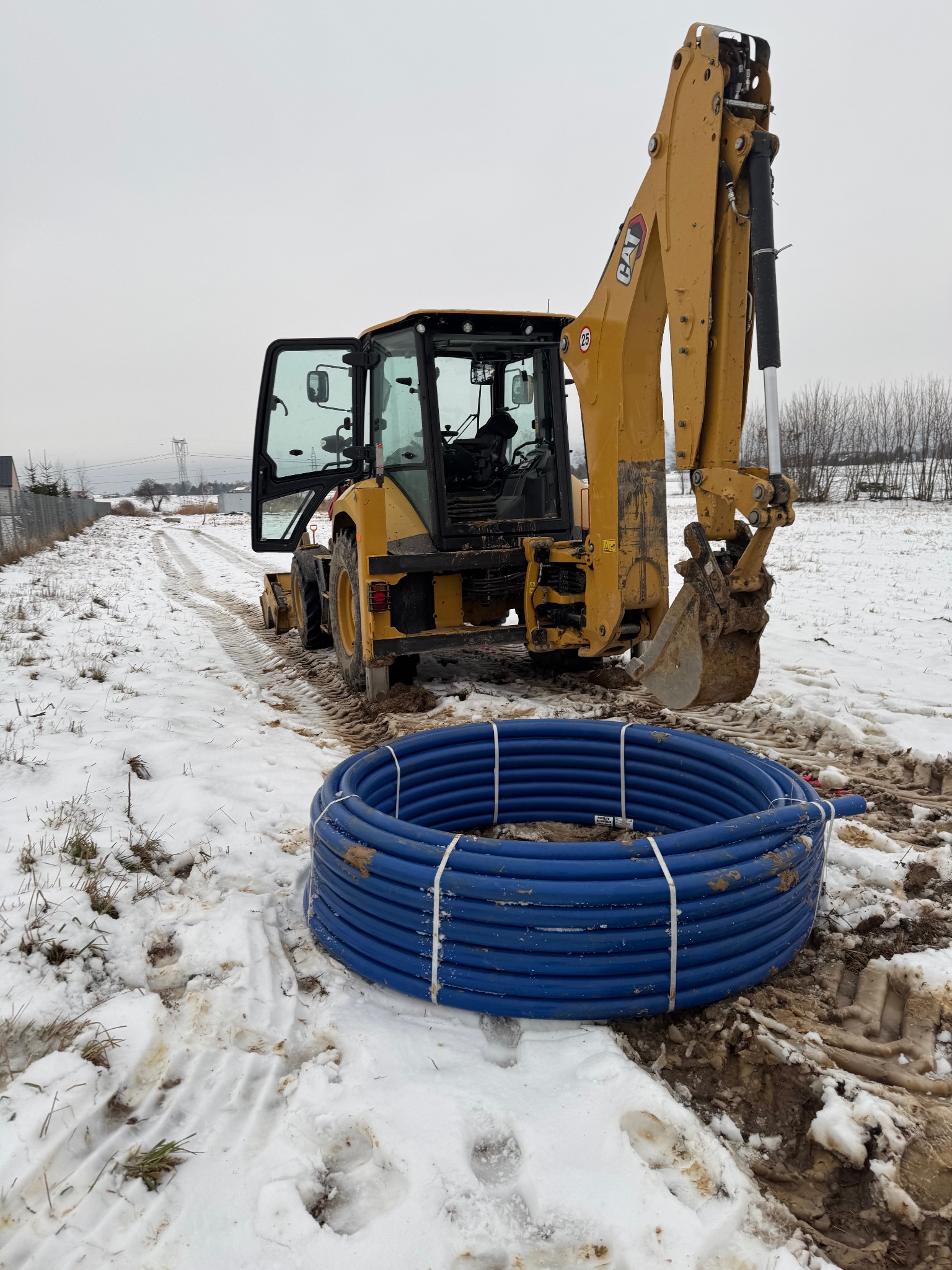 Żółta koparko-ładowarka CAT na zasypanym śniegiem placu budowy, obok zwój niebieskich rur z tworzywa sztucznego, widoczne ślady opon w błocie i śniegu.