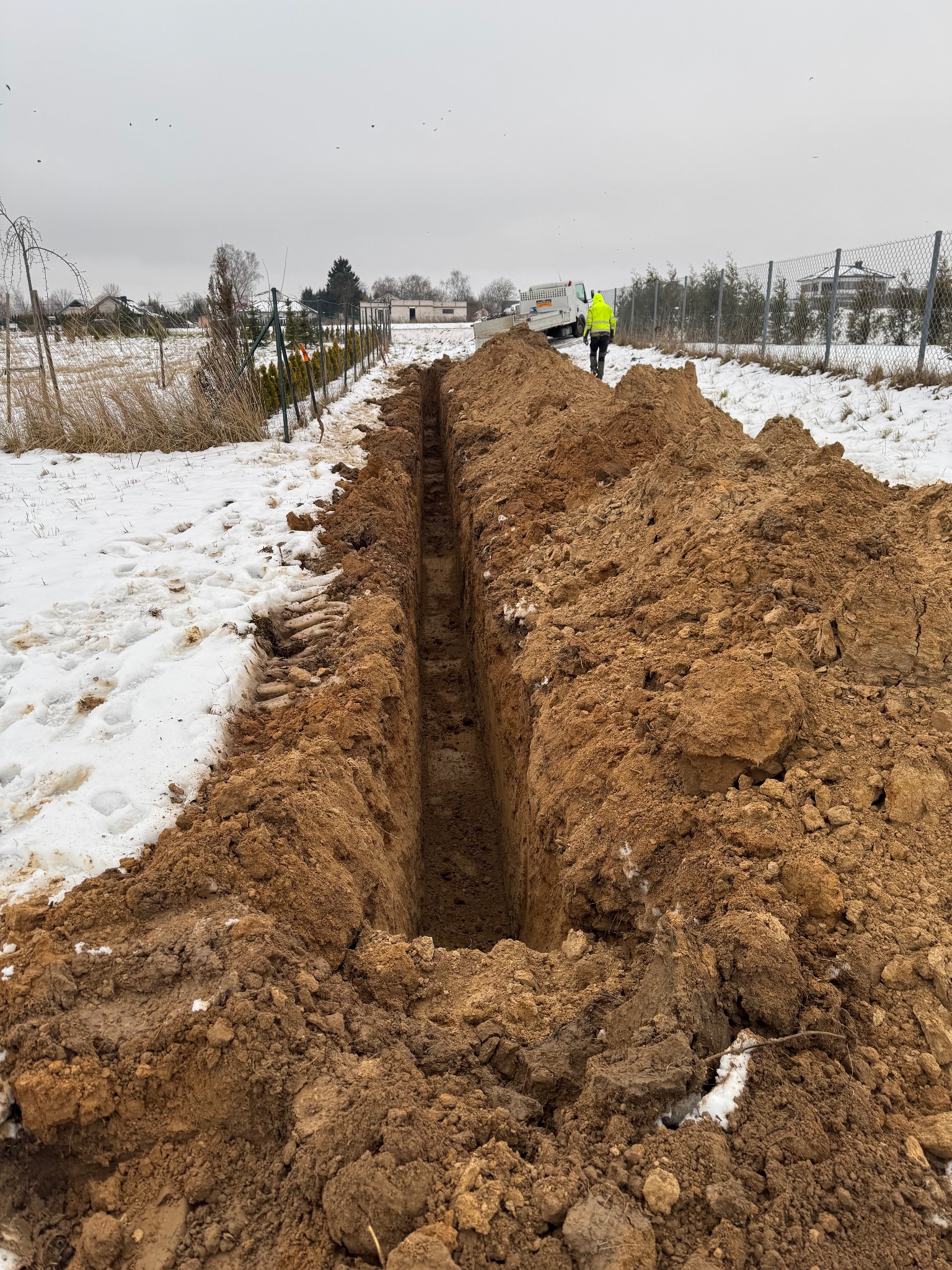 Wąski wykop w ziemi z resztkami śniegu, w tle pracownik w żółtej kamizelce i biały samochód dostawczy na tle pochmurnego nieba i ogrodzenia.