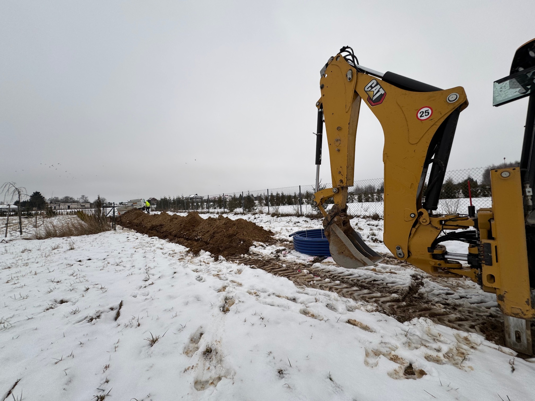 Żółta koparka CAT przy wykopie w zasnieżonym terenie, widoczne niebieskie rury i pracownik w oddali.