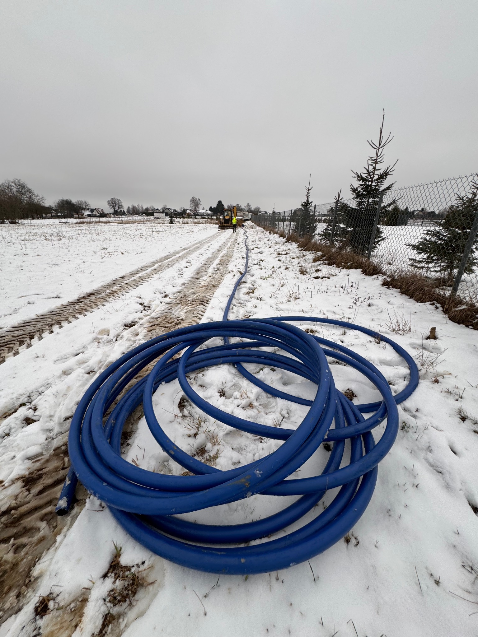 Zwój niebieskiego węża na śniegu, w tle wykop i koparka w zimowej scenerii, widoczne ślady opon na ziemi.